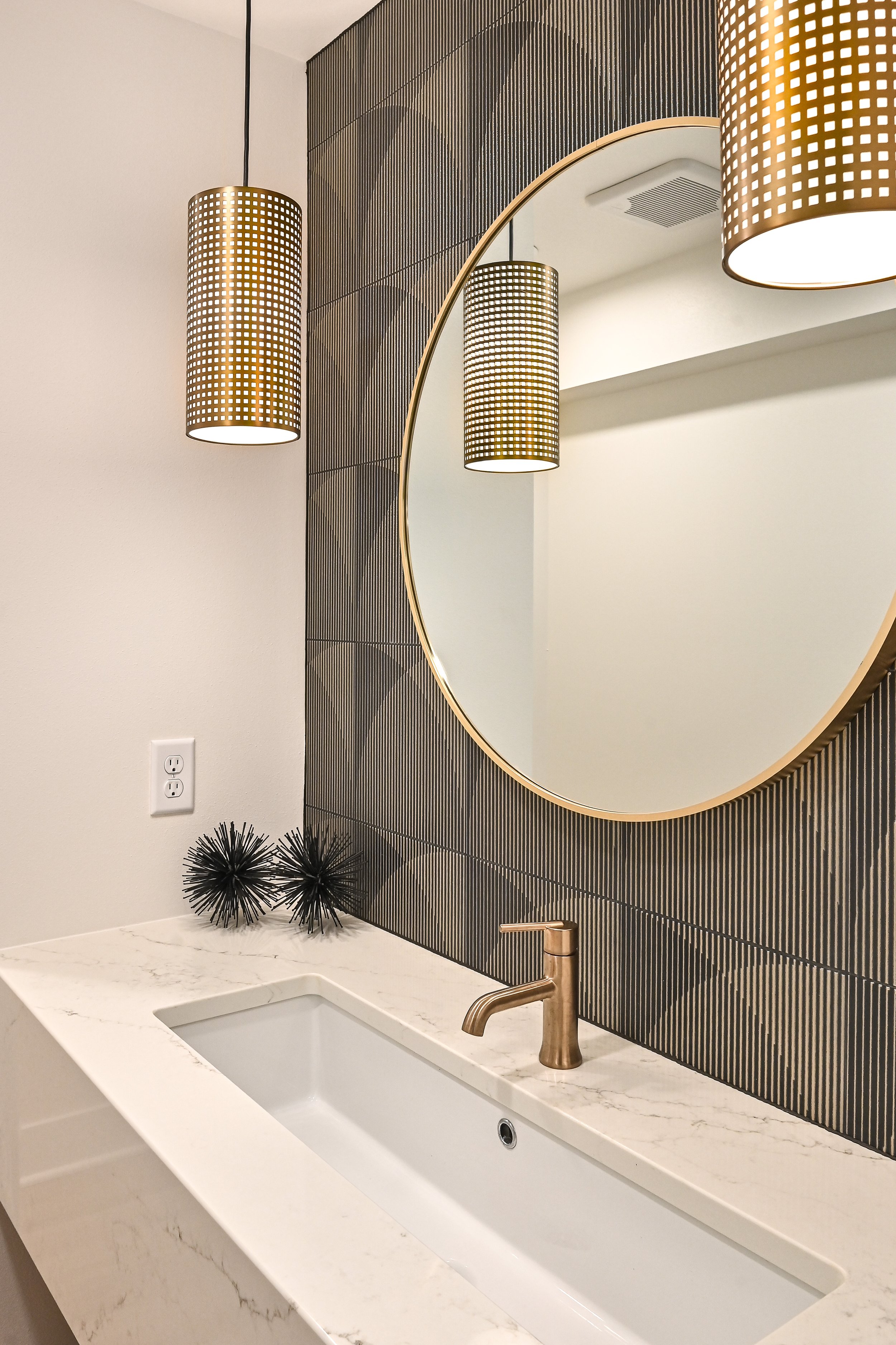 Bathroom vanity with a white marble countertop, gold faucet, large round mirror, black geometric decorations, and pendant lights with a gold grid design.