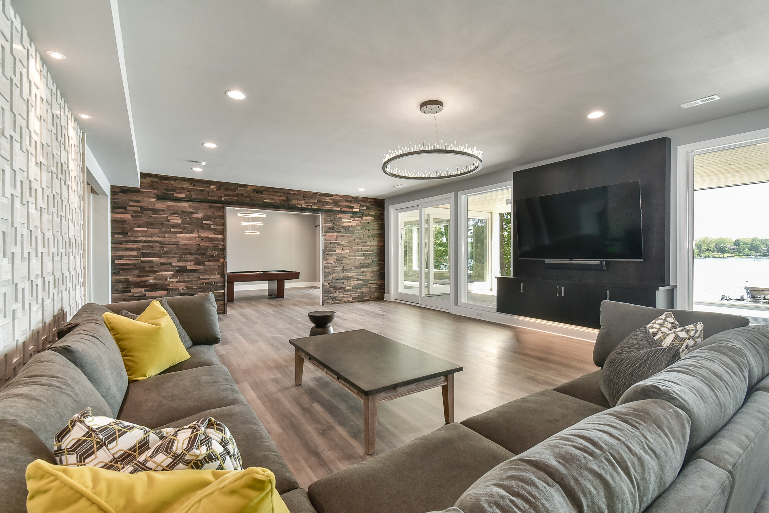 Modern living room with large L-shaped gray sectional sofa, yellow and patterned throw pillows, a wooden coffee table, a black entertainment wall with a flat-screen TV, a circular chandelier, and twin sliding glass doors leading to a porch overlookin