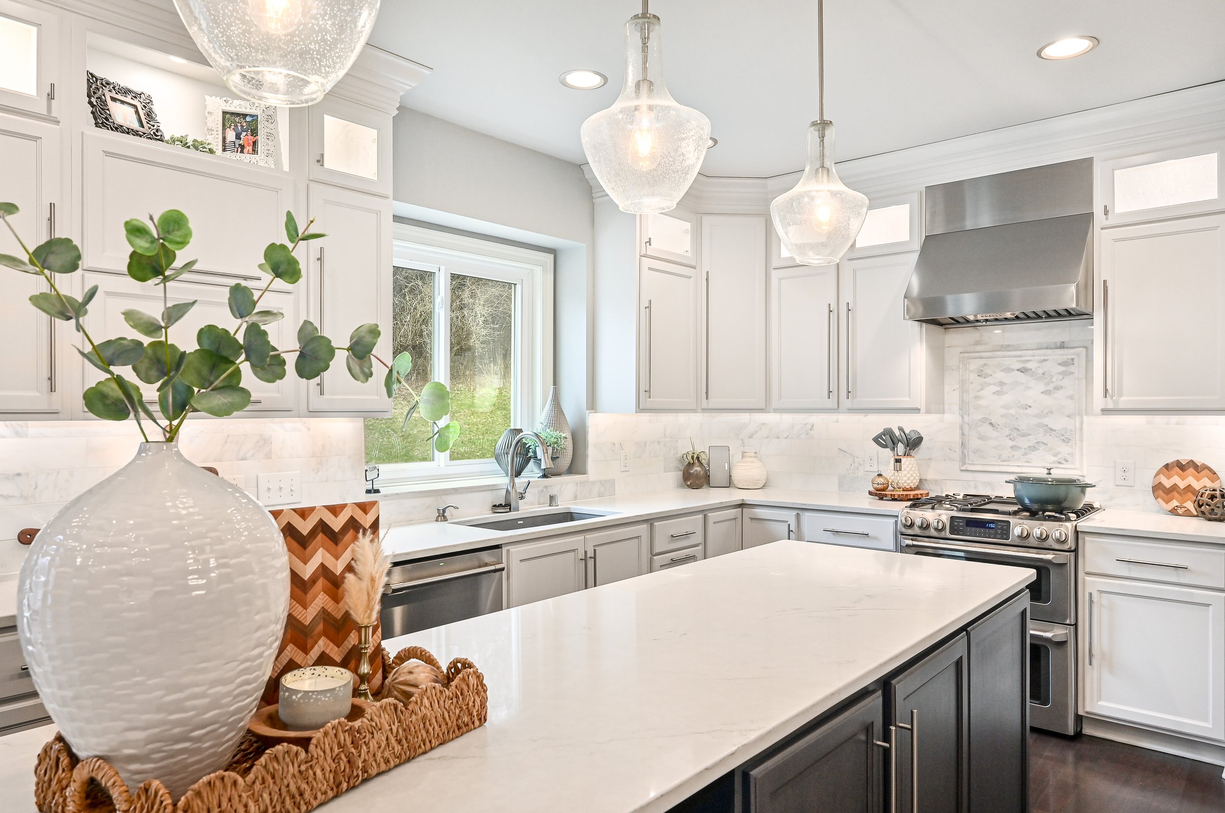 Modern kitchen with white cabinets, gray lower cabinets, marble countertops, stainless steel appliances, and decorative accents, including vases and candles.