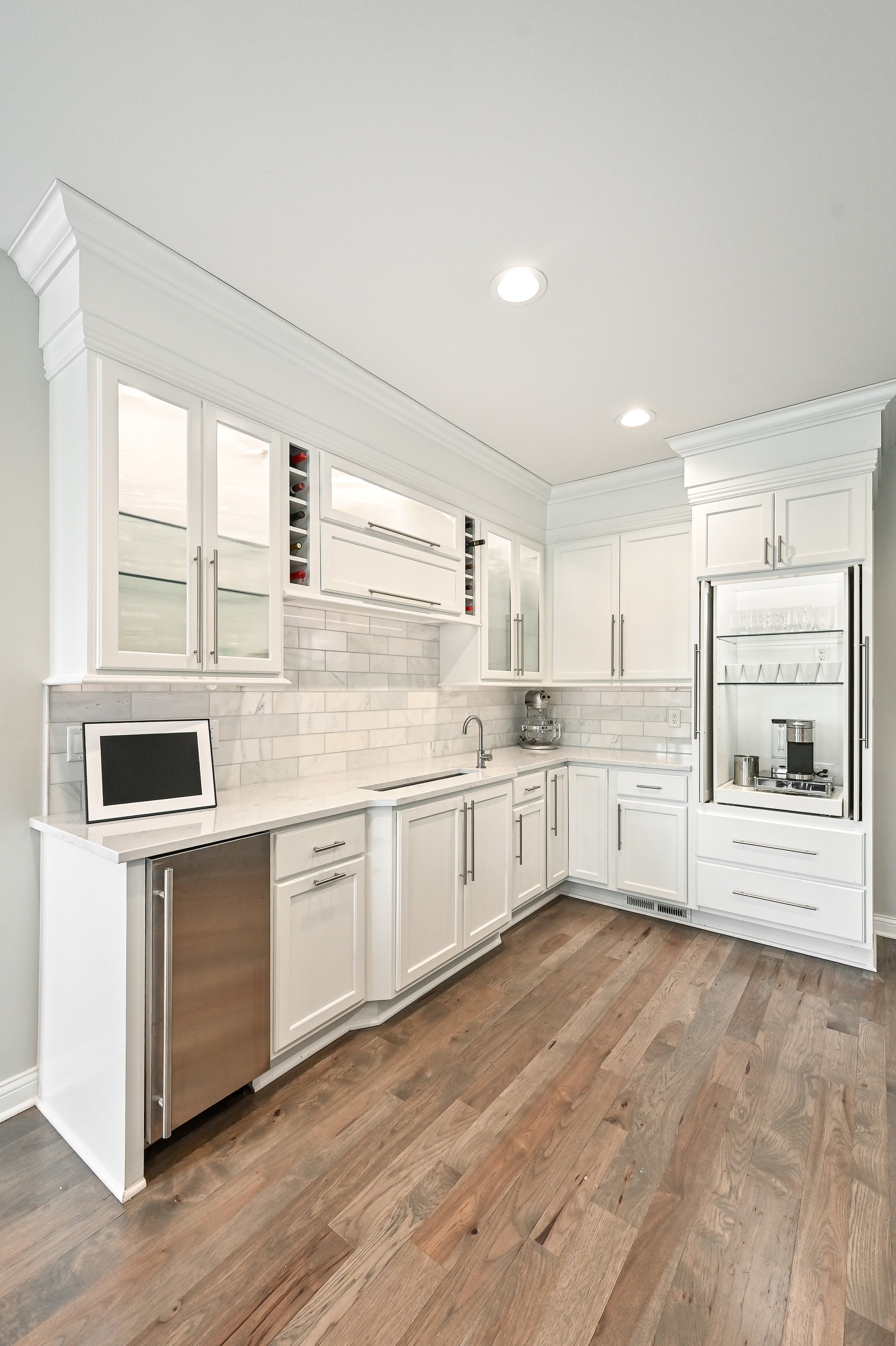 Bright white kitchen with wood flooring, upper glass-front cabinets, and a marble backsplash; includes a small tablet on the counter, a sink, and a coffee station.