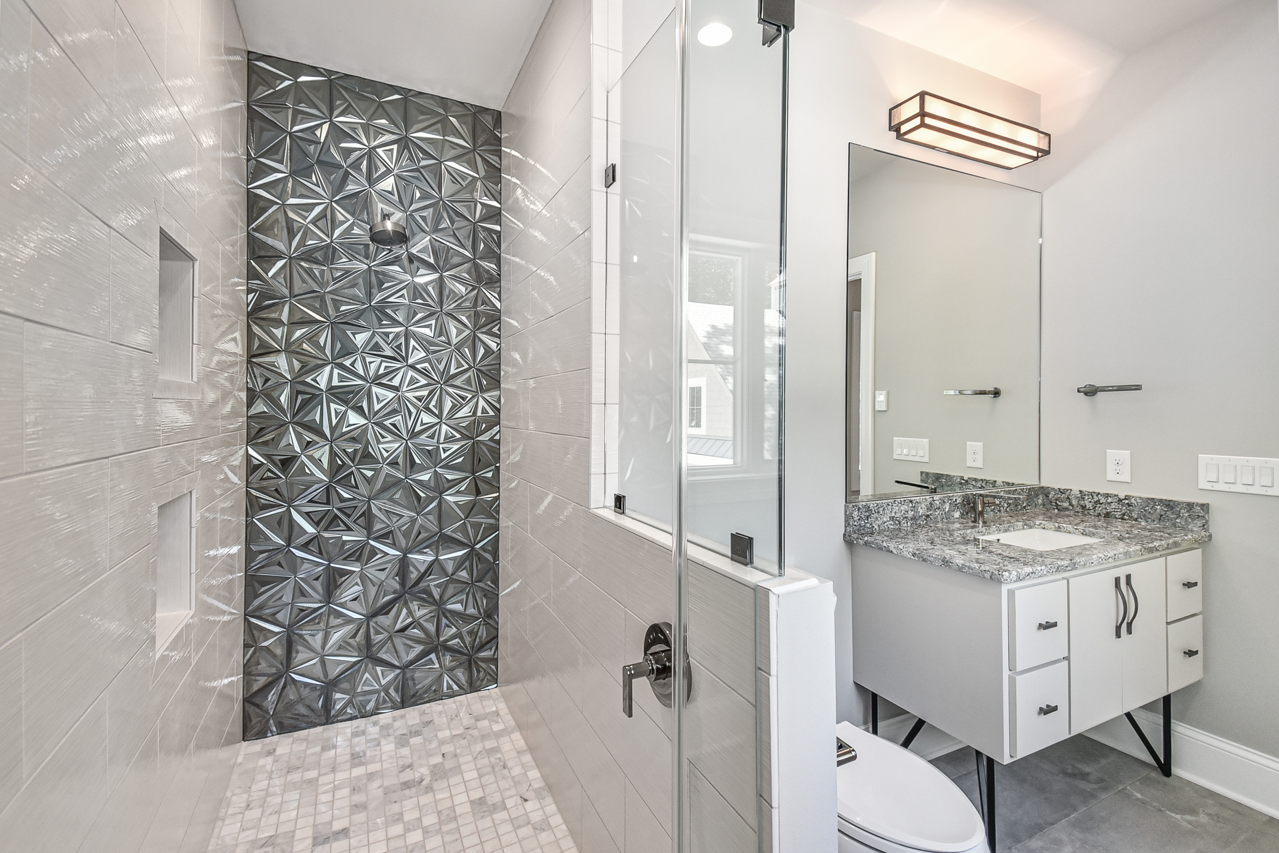 Modern bathroom with a glass-enclosed shower featuring a decorative textured black tile accent wall, and a vanity with a granite countertop, large mirror, and wall-mounted light fixture.