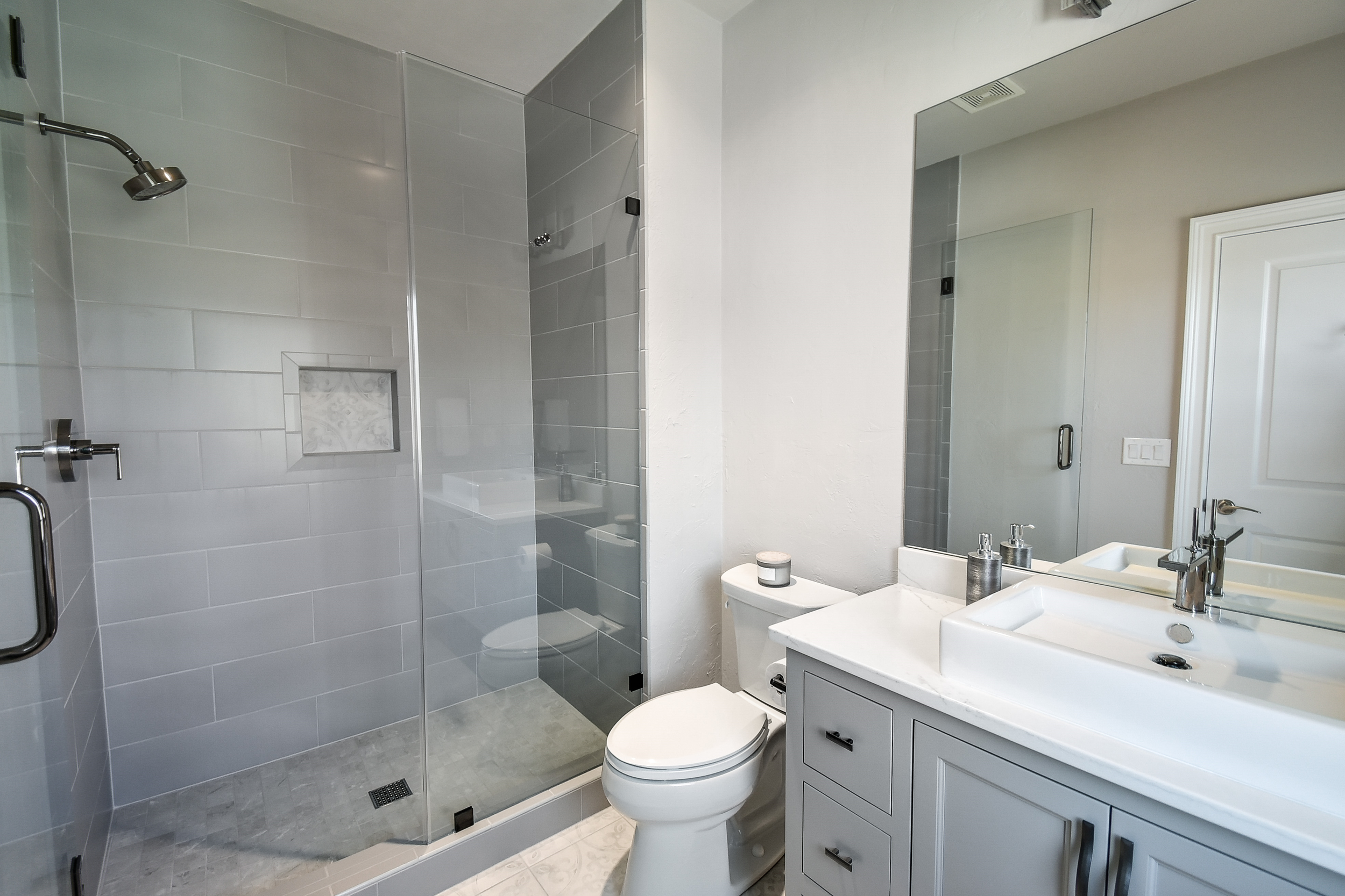Modern bathroom with glass shower enclosure, white toilet, and gray vanity with sink, large mirror, and gray walls.