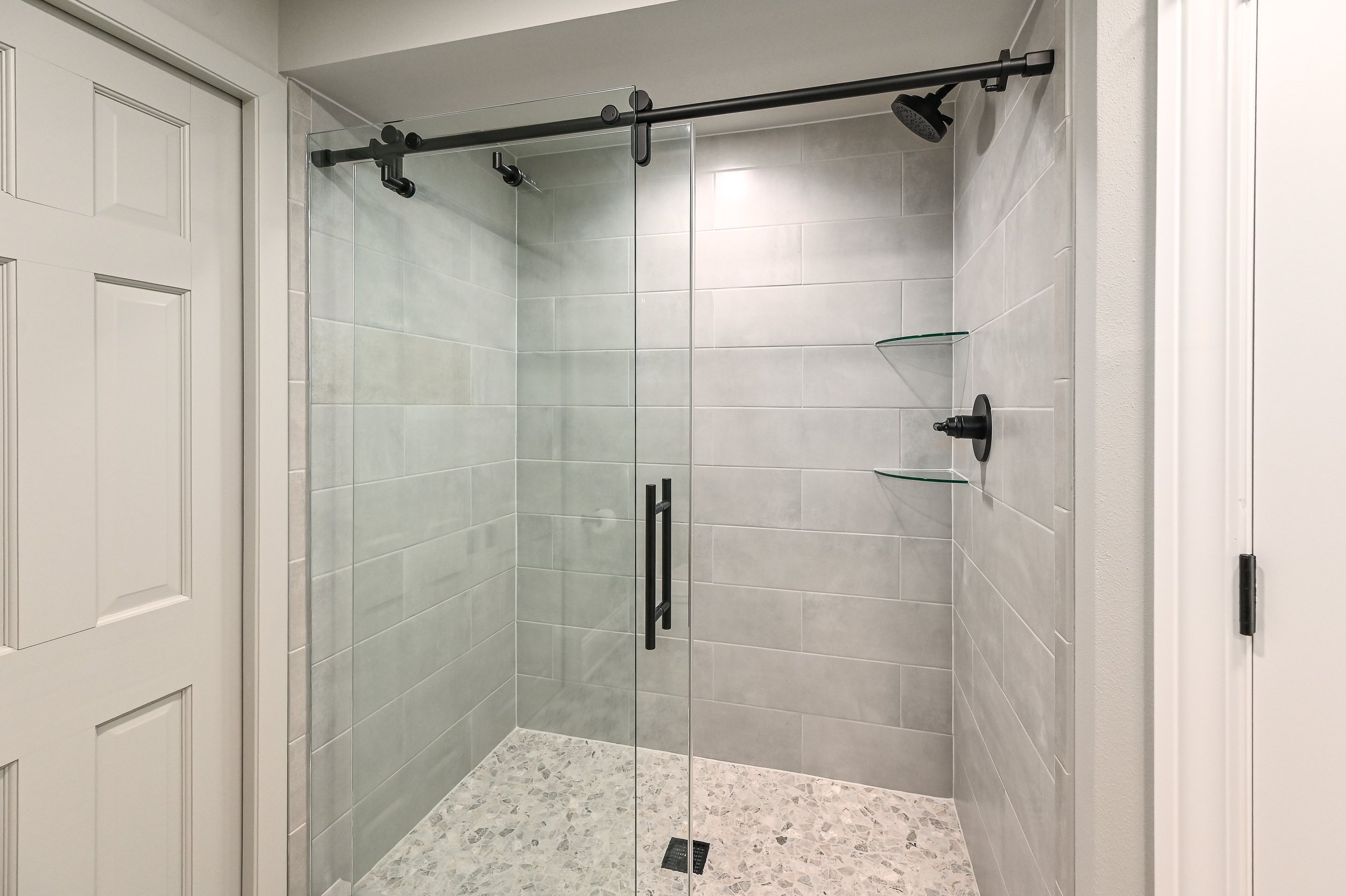 Modern walk-in shower with glass sliding doors, black hardware, gray tiled walls, and a mosaic tile floor.