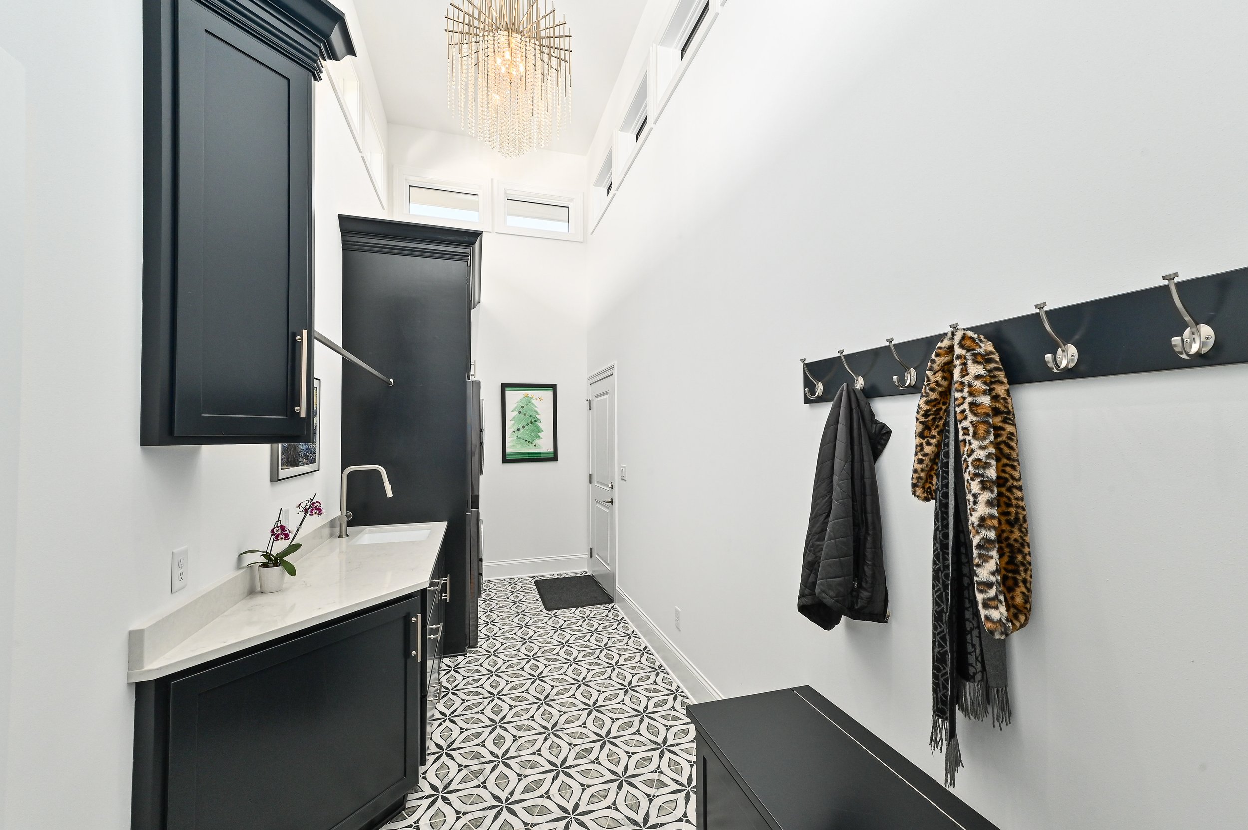 Modern entryway with black cabinets, a wall-mounted coat rack with coats and scarves, patterned floor tiles, a small white sink with a plant, and a large chandelier.