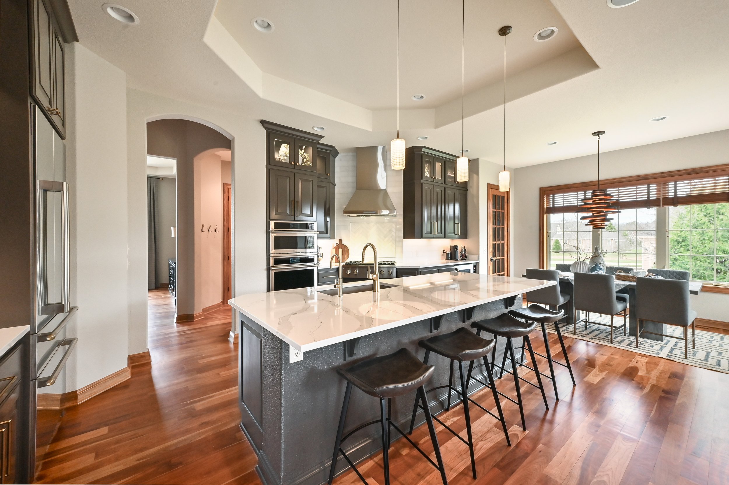 Open-concept kitchen with dark gray cabinets, a large island with marble countertop, and a dining area by a big window showing outdoor greenery.