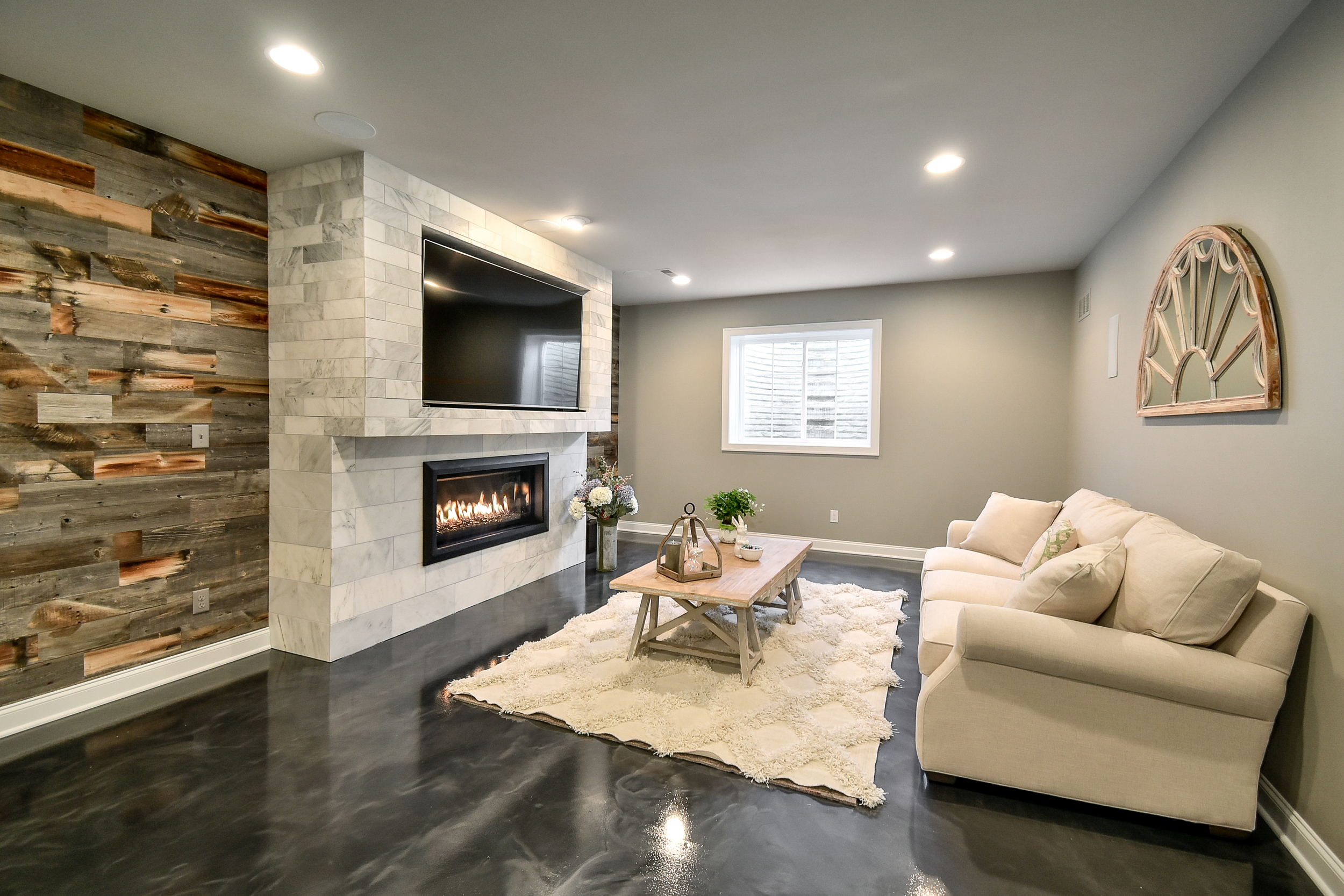 Living room with a beige sofa, a wooden coffee table on a fluffy white rug, a wall-mounted TV above a modern fireplace, a window with white blinds, a decorative wall piece, and a wooden accent wall.