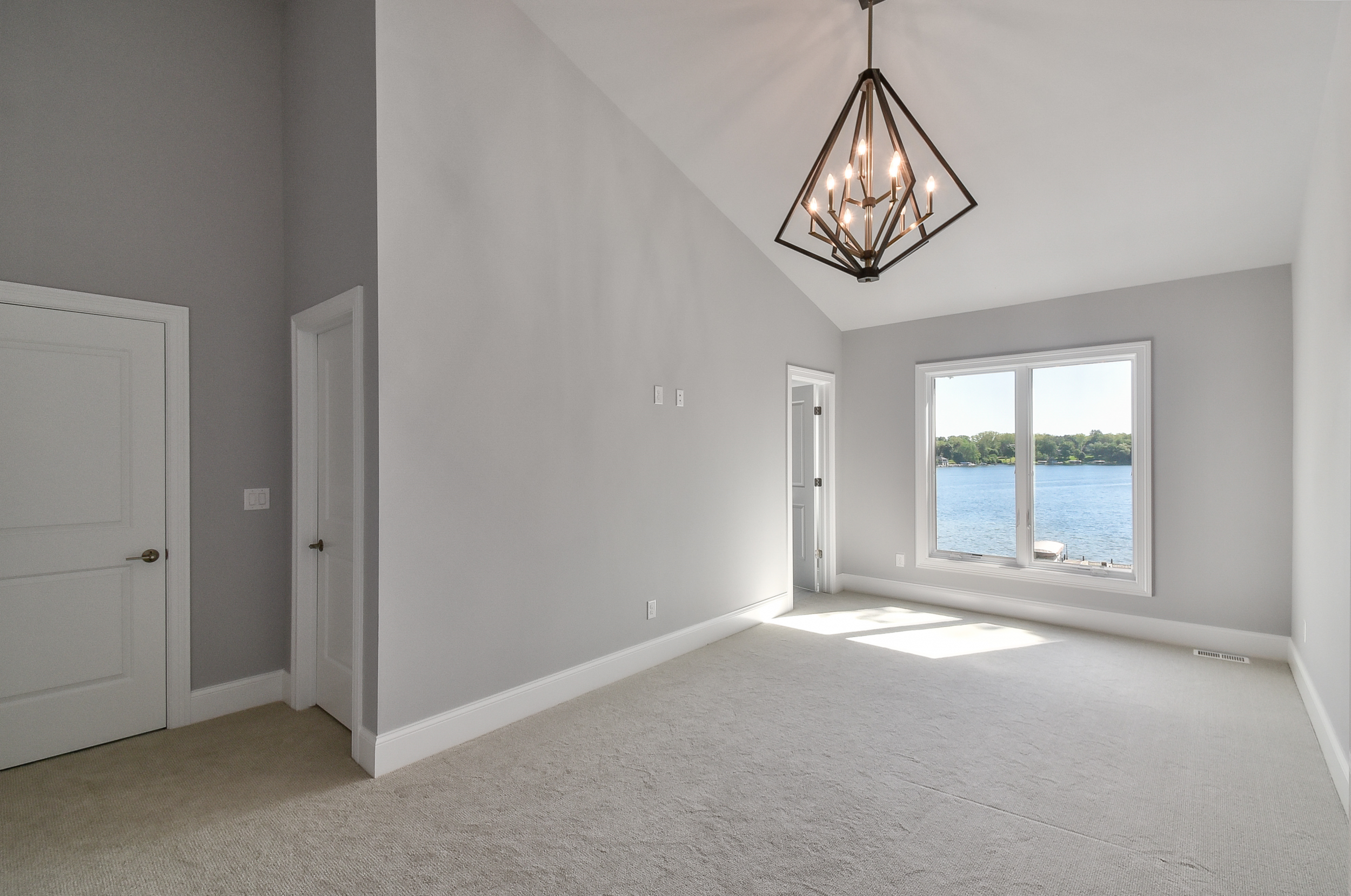 Empty room with light gray walls, beige carpet, large window with a view of a lake, and a geometric chandelier hanging from the ceiling.