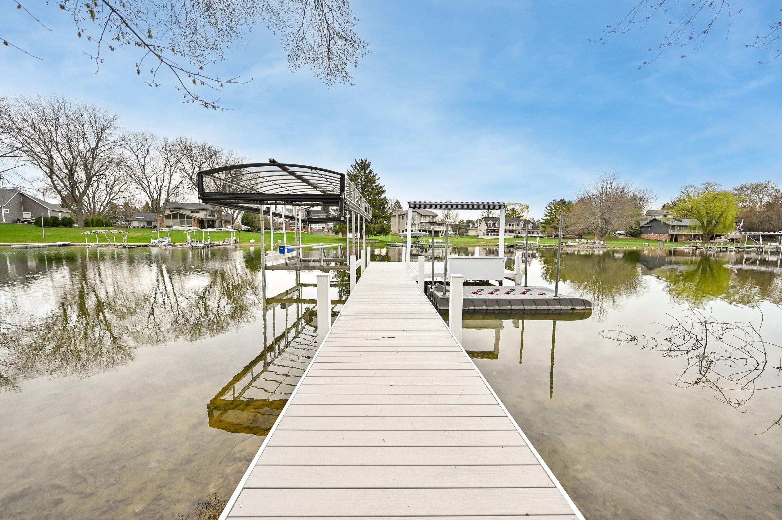 A white wooden dock extending into a calm lake with houses and trees in the background, under a partly cloudy sky.