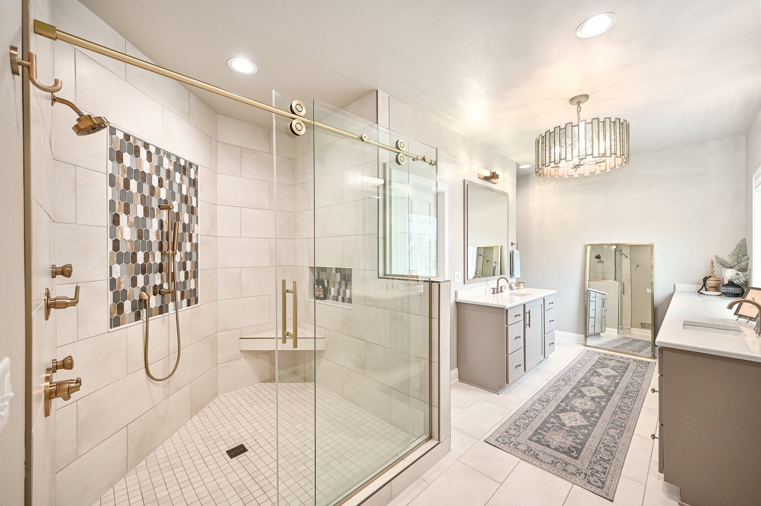 Modern bathroom with a glass-enclosed shower featuring a decorative mosaic tile accent, a large vanity with double sinks, a framed mirror, a decorative rug, and a chandelier