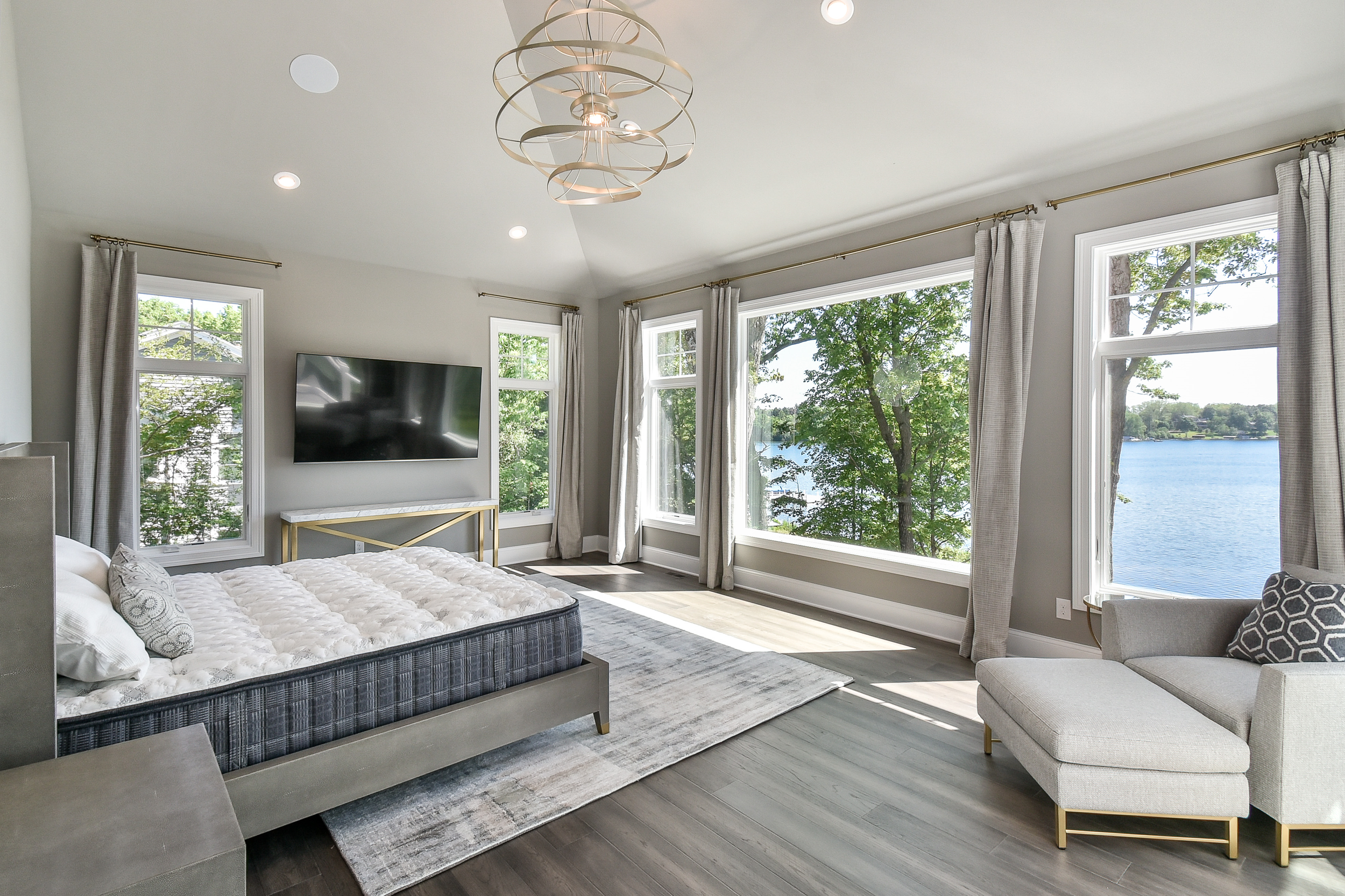 A bright, modern bedroom with large windows overlooking a lake, featuring a bed with a gray headboard, a gray chaise lounge with pillows, a wall-mounted TV, a small gold and white console table, and a patterned area rug.