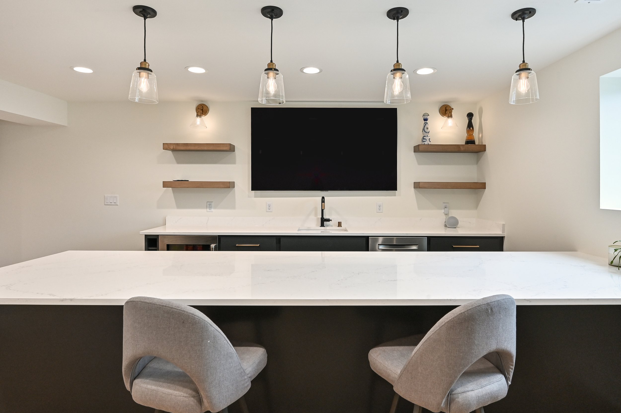 Modern kitchen with white countertops, two upholstered bar stools, a black cabinet, and floating wooden shelves. There are ceilings lights and decorative items on the shelves, with an empty black picture frame or chalkboard on the wall.