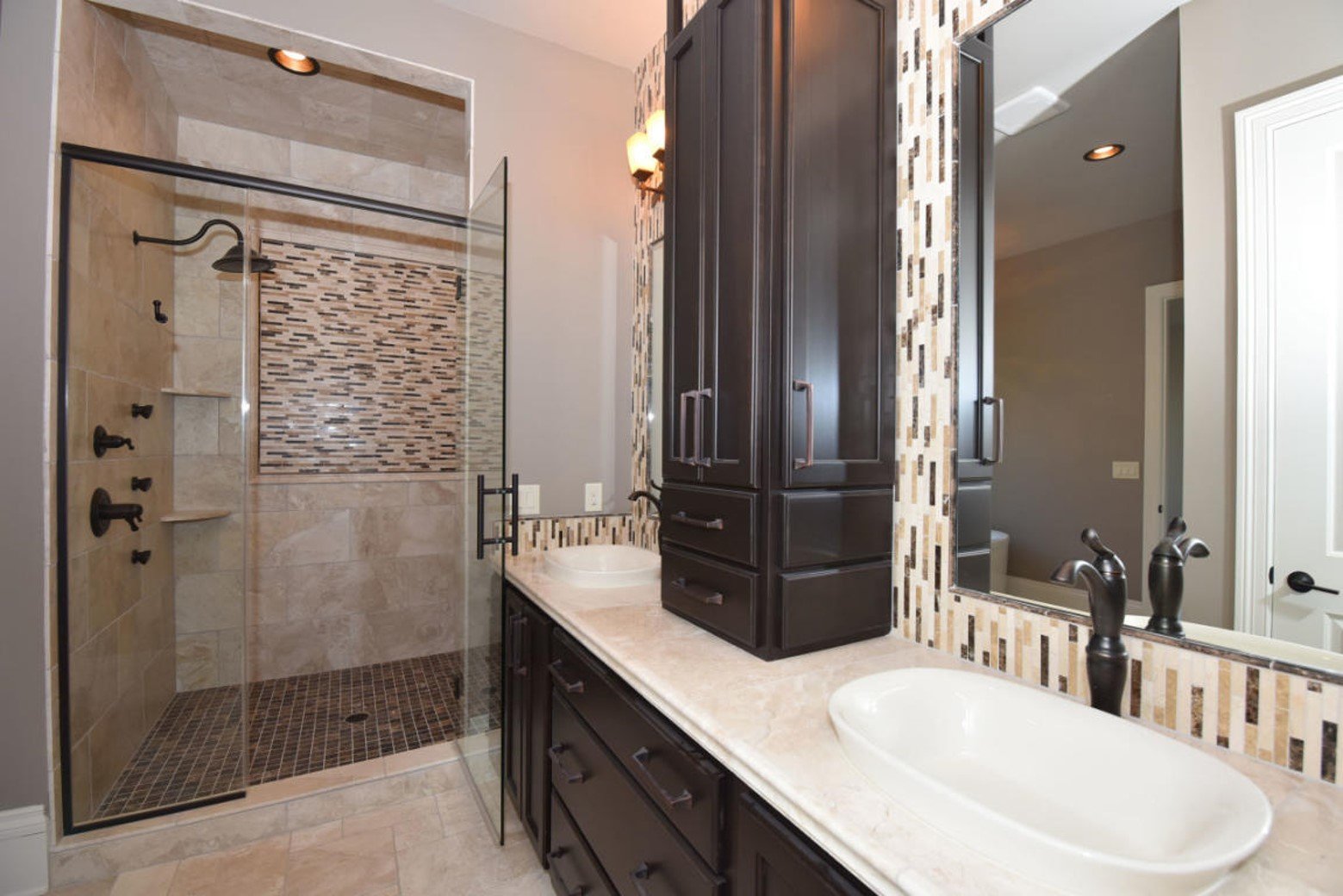Modern bathroom featuring a glass-enclosed walk-in shower with beige and mosaic tile accents, a white oval sink with black faucet, dark wood cabinetry, and a decorative tile border behind the mirror.