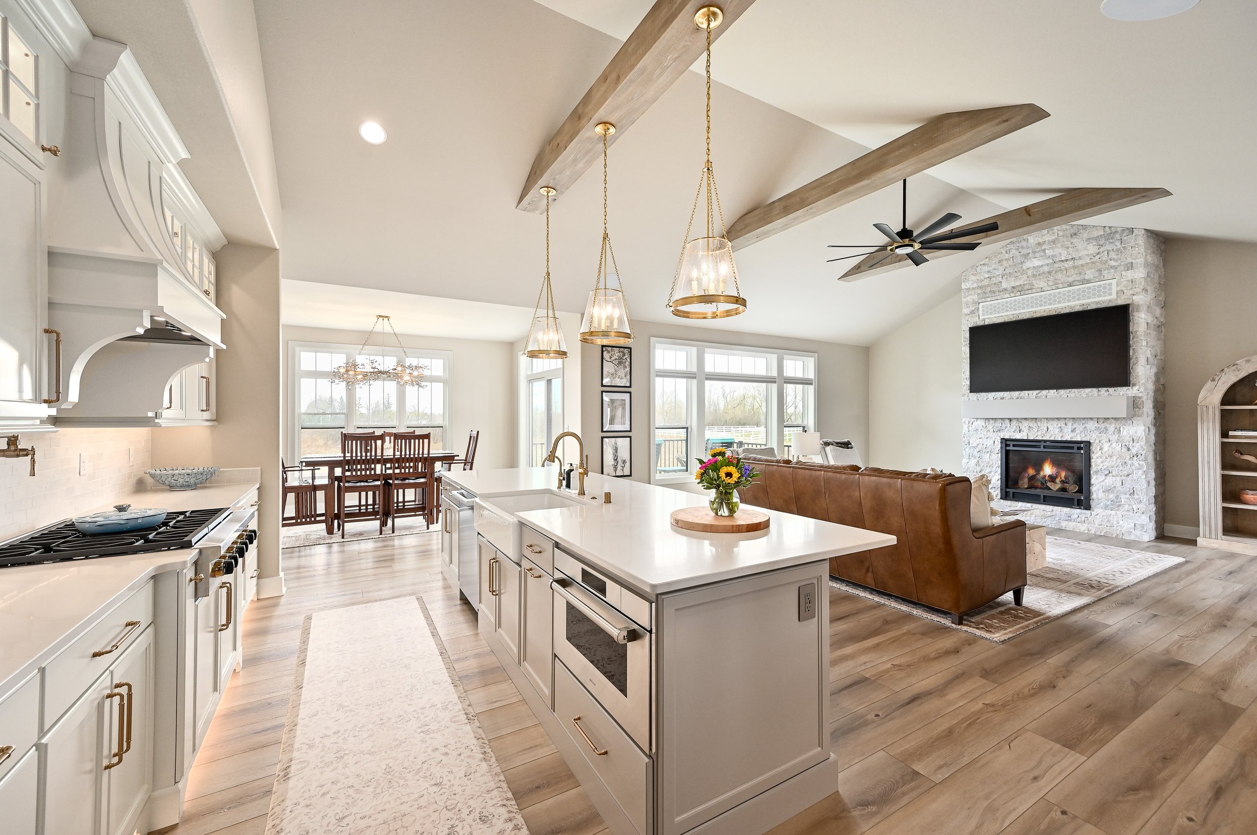 Open-concept living room and kitchen with white cabinets, a kitchen island with a white countertop and gold hardware, a brown leather sofa facing a white brick fireplace with a mounted TV, and a dining area with wooden chairs and a chandelier, all wi