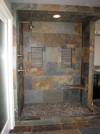 A spacious walk-in shower with multicolored natural stone tiles on the walls, two built-in shelves, and pebble stone flooring. A recessed ceiling light illuminates the shower area.