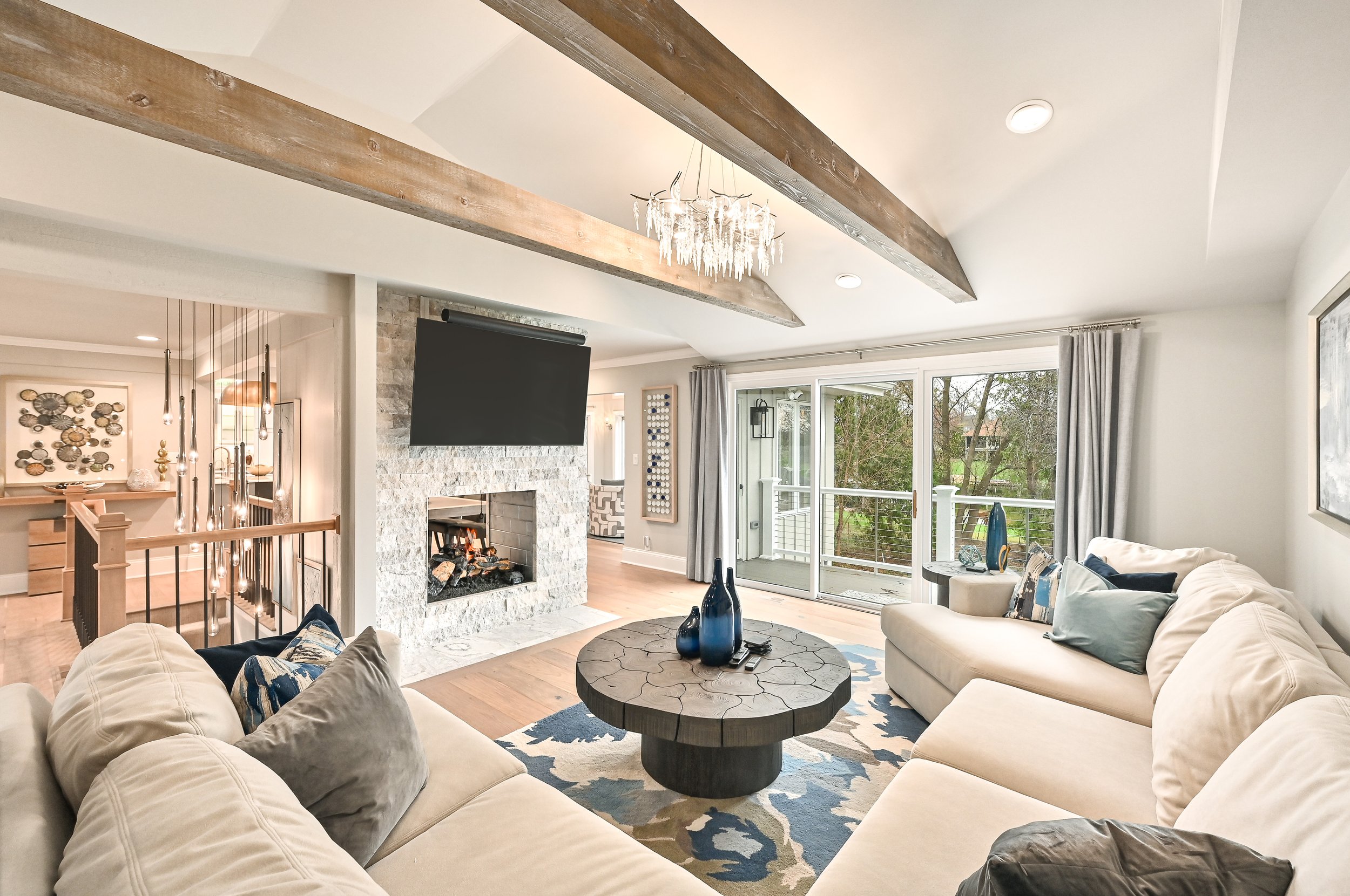 Bright living room with white sofas, a round wooden coffee table, and a white stone fireplace with a mounted TV. Large sliding glass doors lead to a balcony with greenery outside. Wooden beams and a chandelier hang from the ceiling.