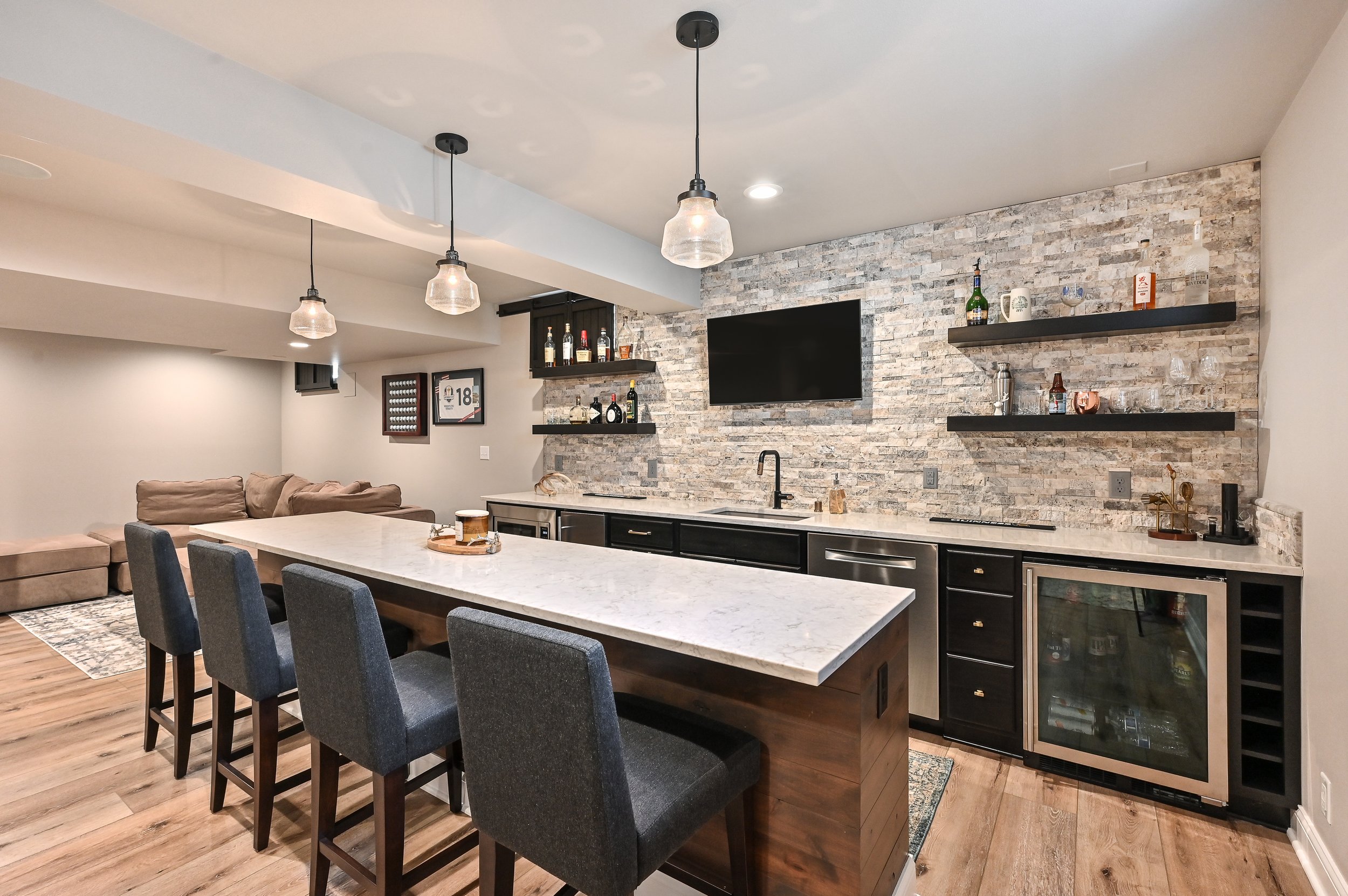 Modern kitchen and living area with a marble island, pendant lights, open shelves on a brick wall with bottles and glassware, and a TV mounted on the wall.