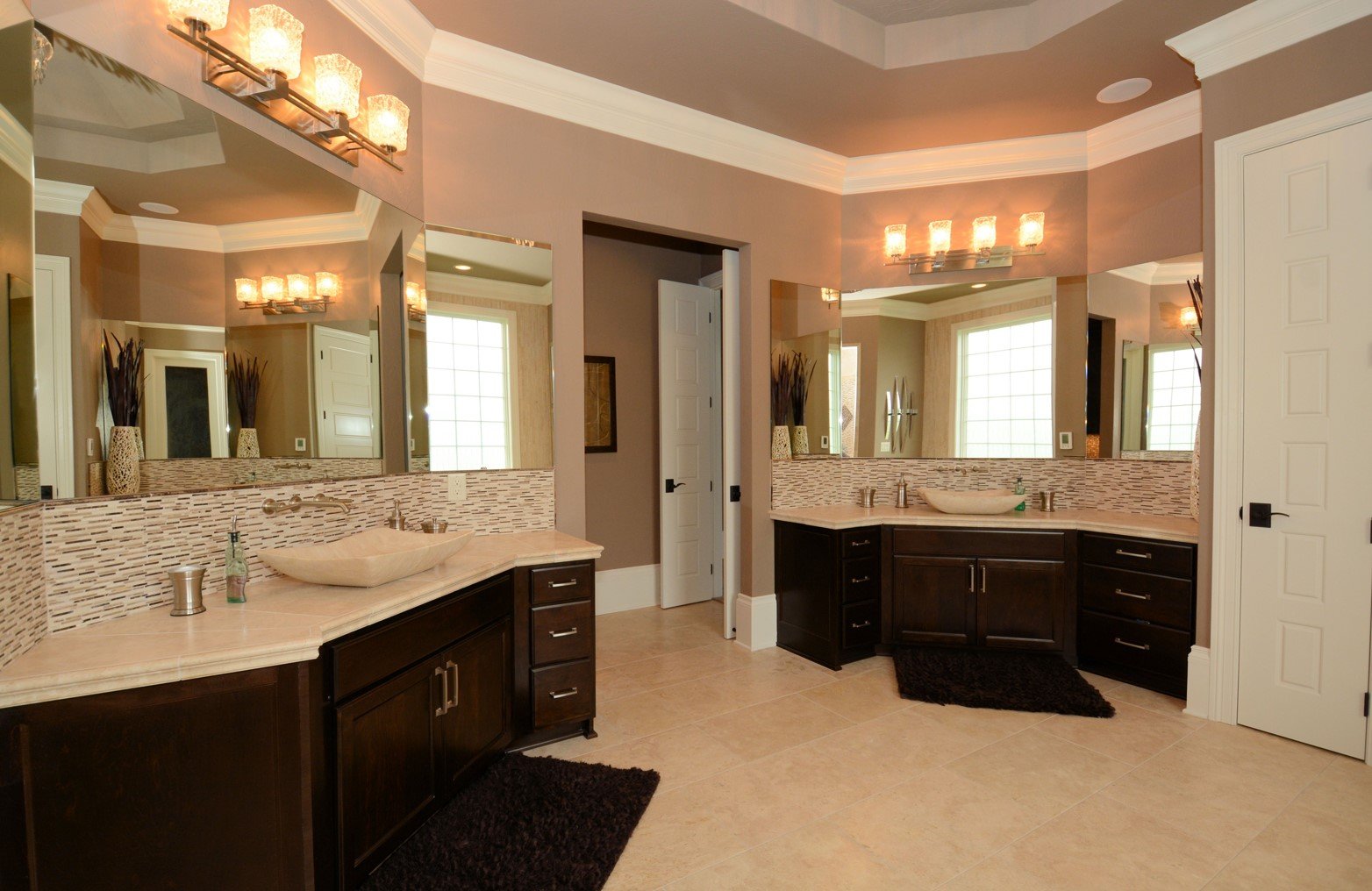 A spacious bathroom with dual vanities featuring dark wood cabinets, marble countertops, and rectangular vessel sinks. Large mirrors above the vanities, modern wall-mounted light fixtures, and decorative vases with tall dried branches. The bathroom h