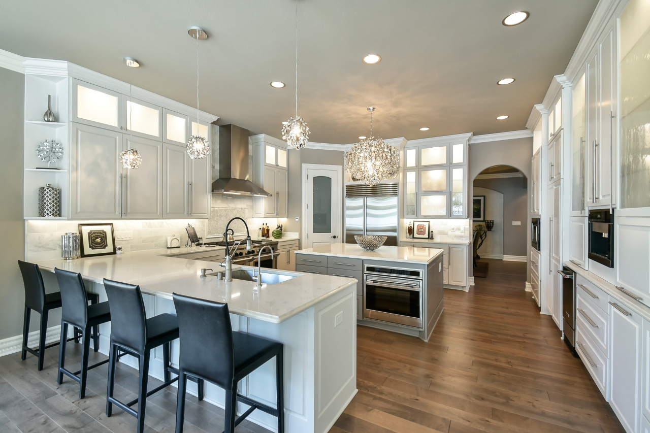 Modern kitchen with white cabinets, an island with a gray base, black bar stools, and stainless steel appliances, including a double oven and a large refrigerator. Decorative lighting fixtures hang from the ceiling.