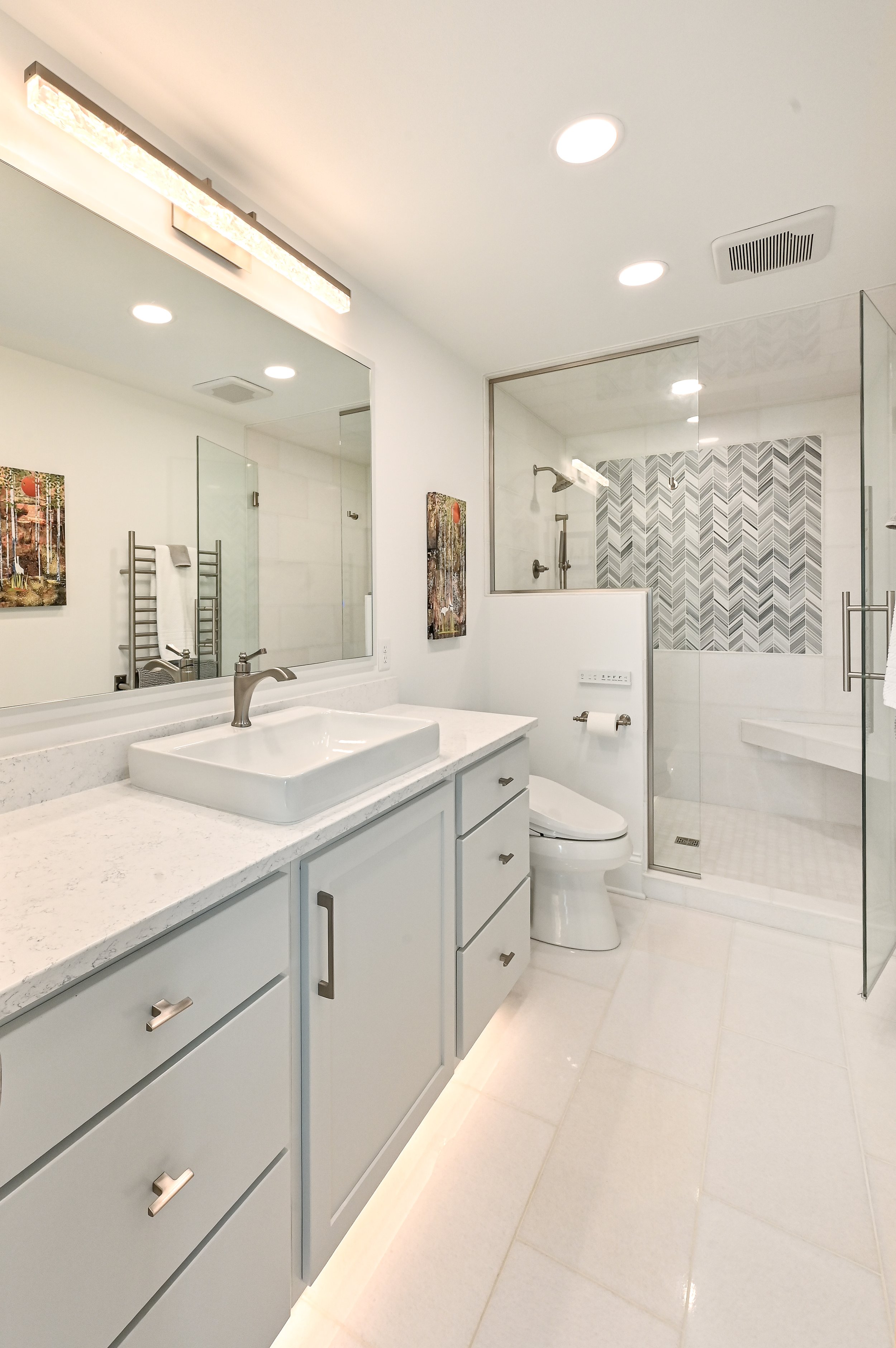 Modern white bathroom with a vanity, vessel sink, large mirror, and a walk-in shower with glass enclosure and gray patterned tiles.