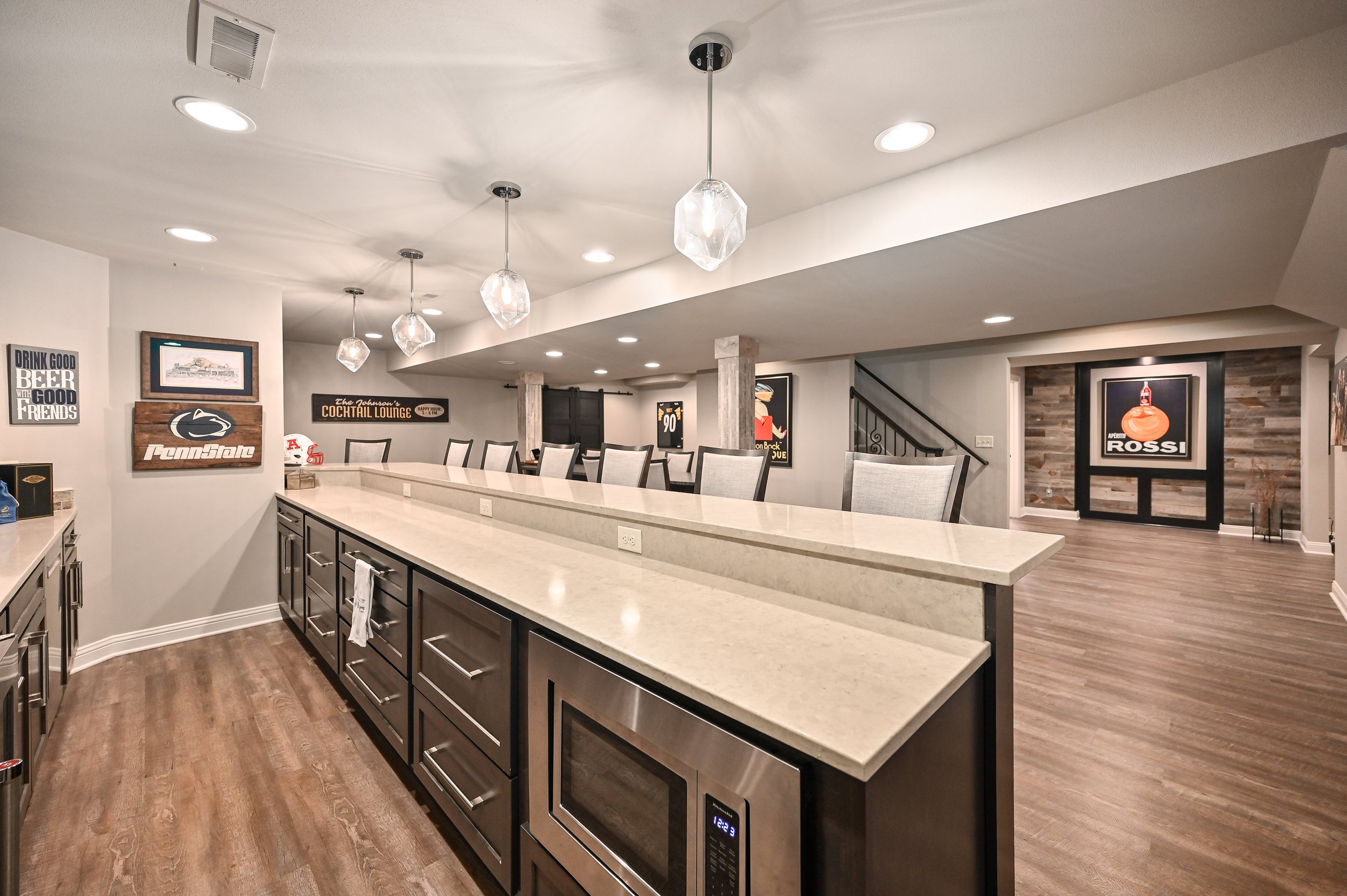 Modern kitchen and dining area with a long beige countertop, dark cabinetry, three pendant lights hanging from the ceiling, and wall decor including Penn State and Rossi signs.