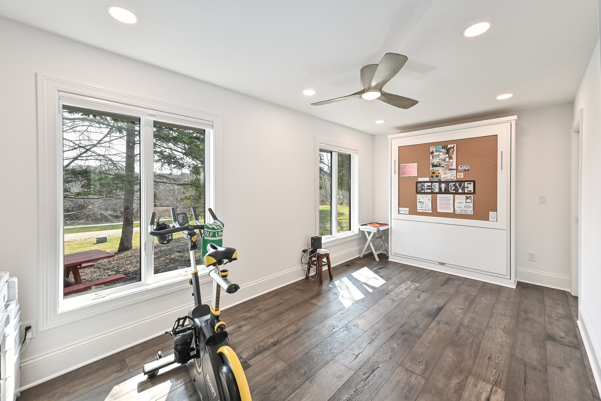 Room with two large windows, exercise bike in the foreground, corkboard with notes and decorations on the wall, and wooden flooring.