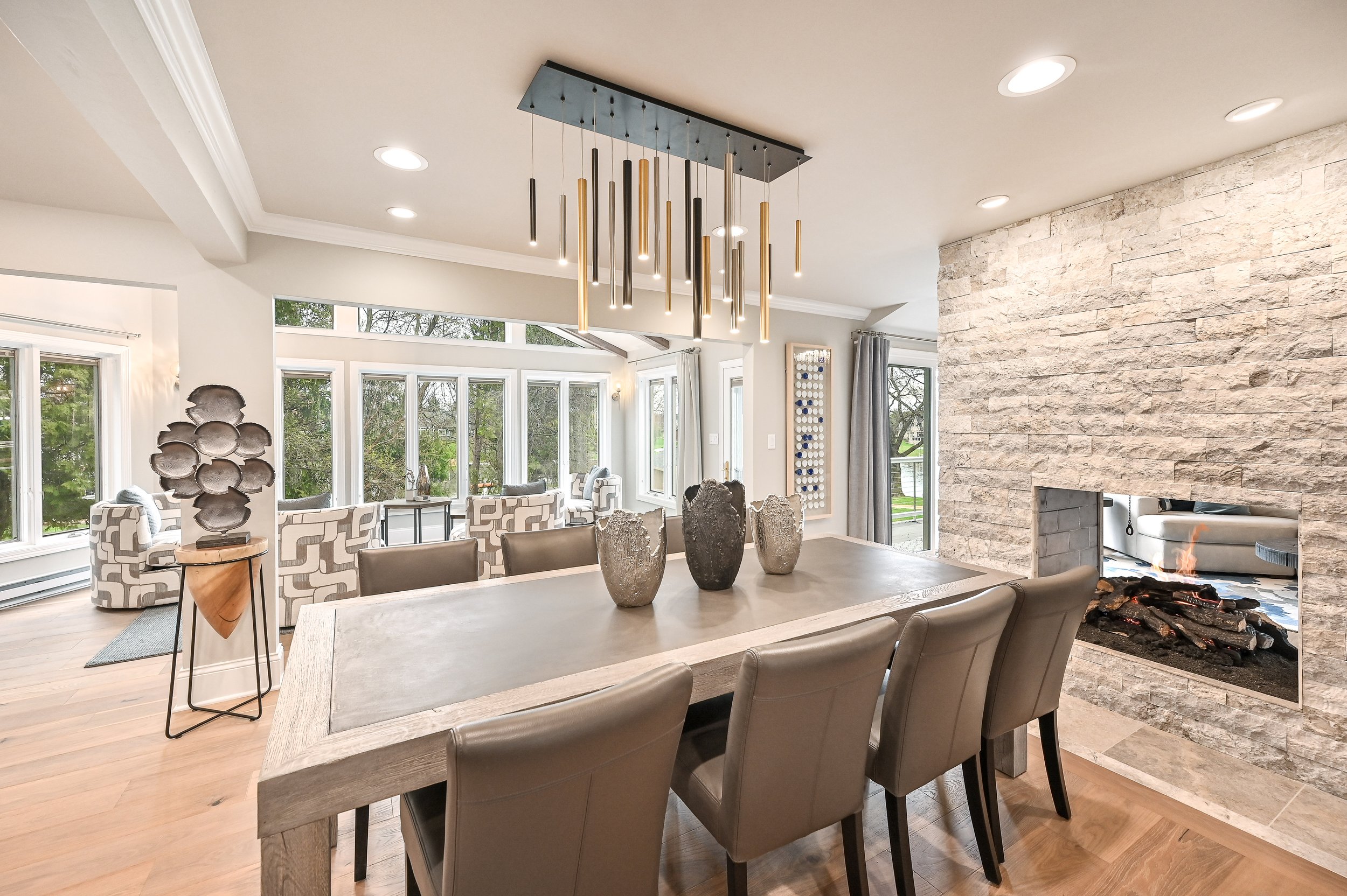 Modern dining room with a large wooden table, six beige chairs, and decorative vases. In the background are sunlit sitting area with patterned armchairs by a bay window, and a stone fireplace with a fire.