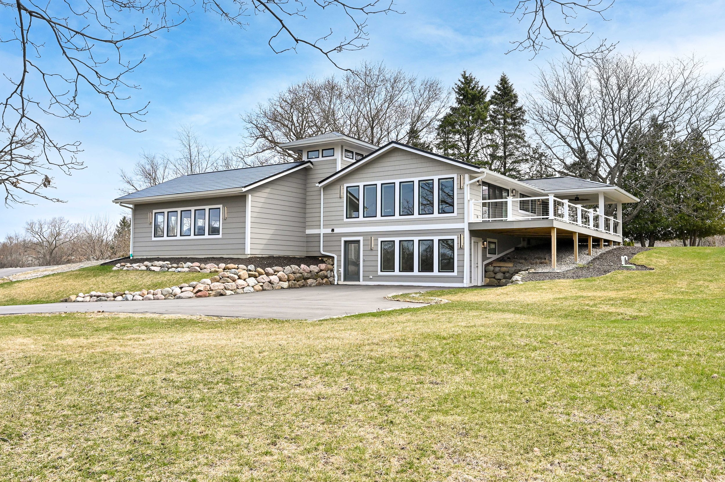 Modern two-story house with gray siding, large windows, a deck, and a landscaped yard with rocks and grass.