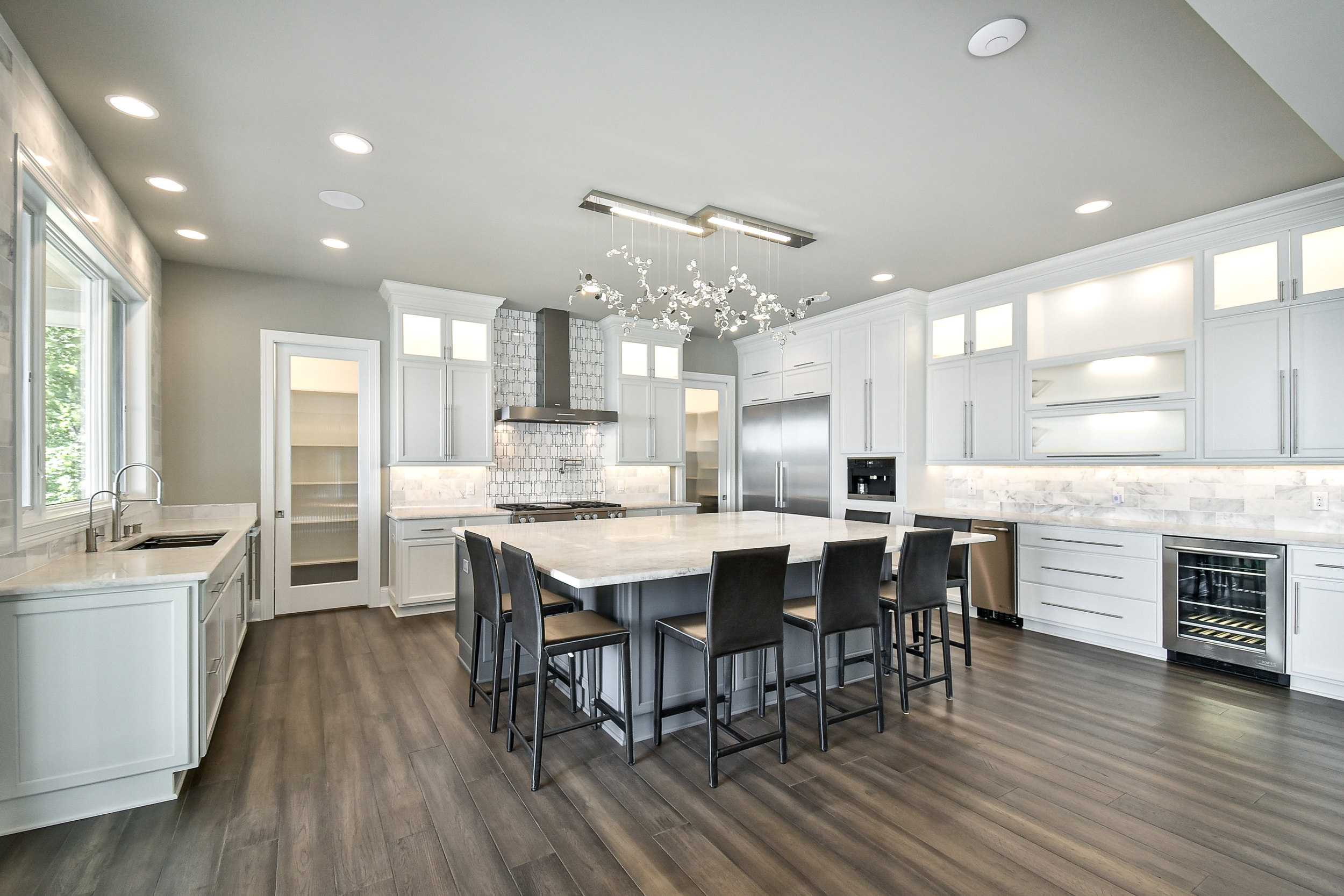 A modern kitchen with white cabinets, a large central island with black chairs, stainless steel appliances, and hardwood floors.