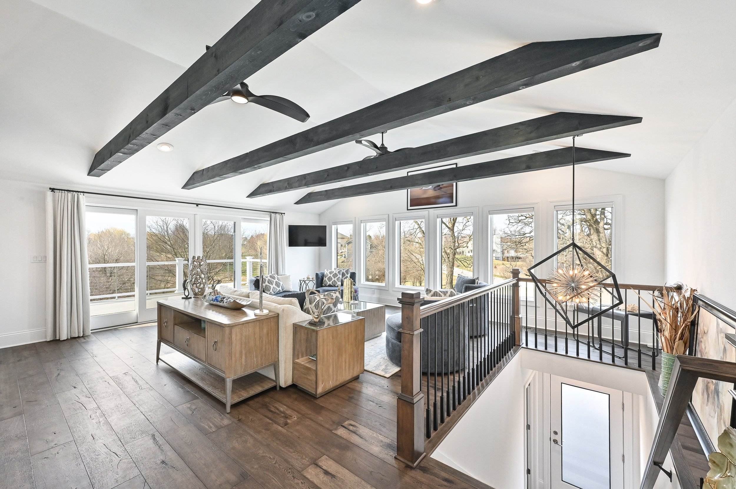 Living room with white walls, large windows, and wooden ceiling beams. It features a sitting area with a beige sofa, gray chairs, and a wooden console table. The room opens to a balcony or patio outside.