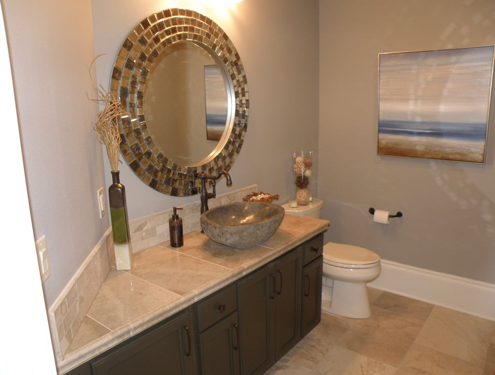 A bathroom with a granite countertop, a decorative vessel sink, a large round mirror, a vase with dried branches, and a painting on the wall.