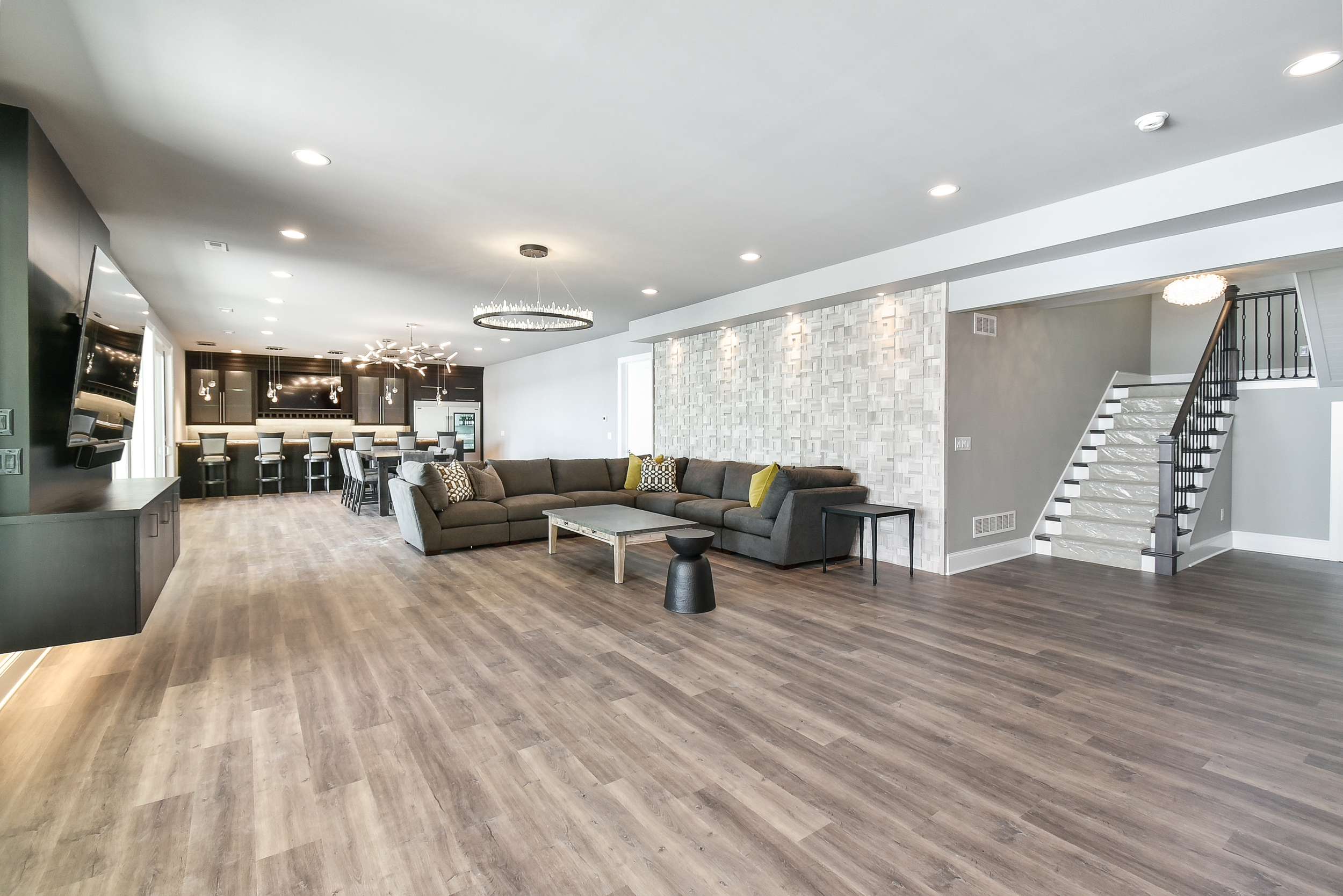 Spacious modern living room with gray sectional sofa, white textured accent wall, large flat-screen TV, and hardwood flooring, open to a dining and kitchen area.