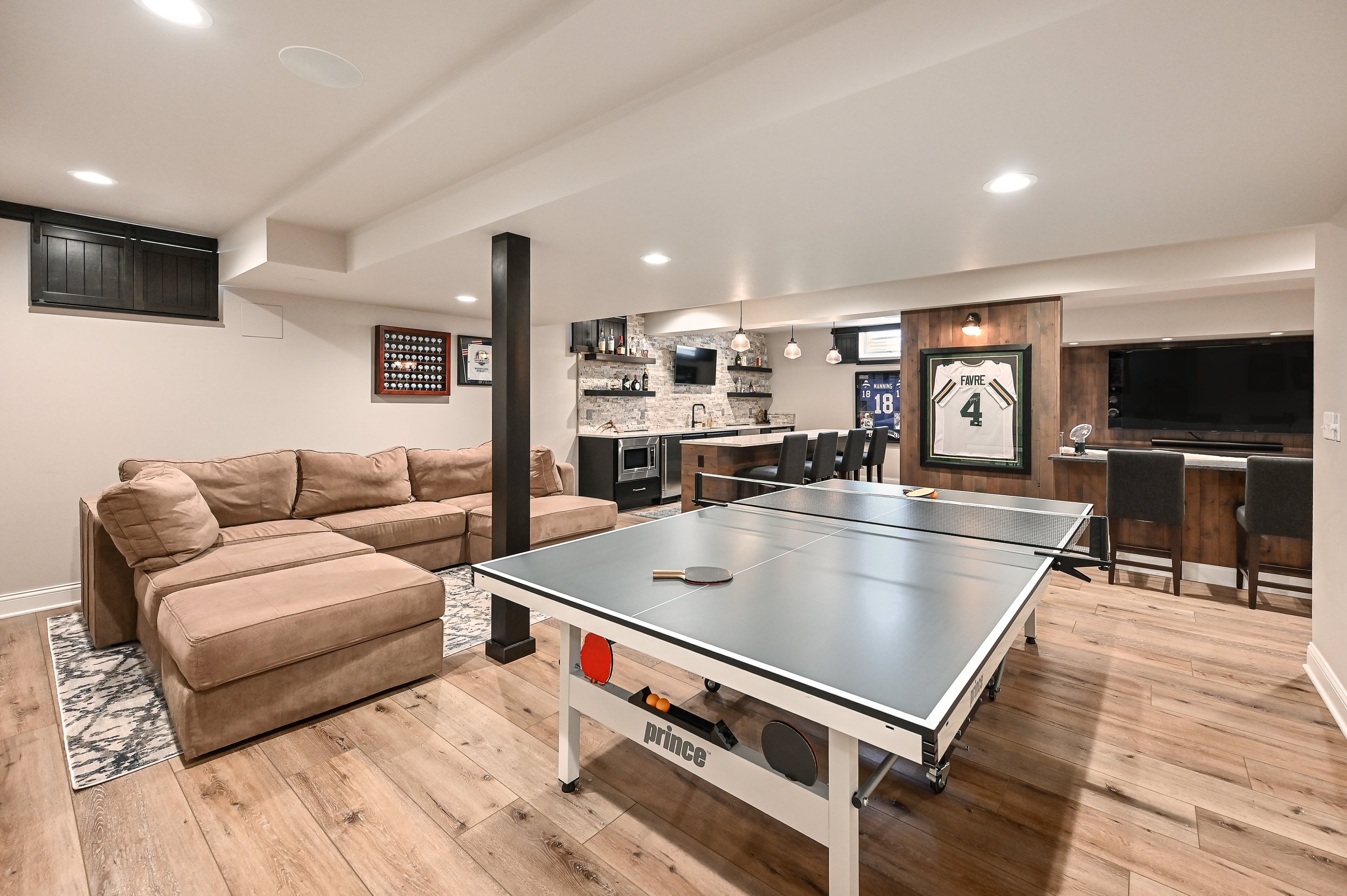 Basement with ping pong table, beige sectional sofa, bar area with four chairs, framed sports jersey, flat-screen TV, and sports memorabilia.