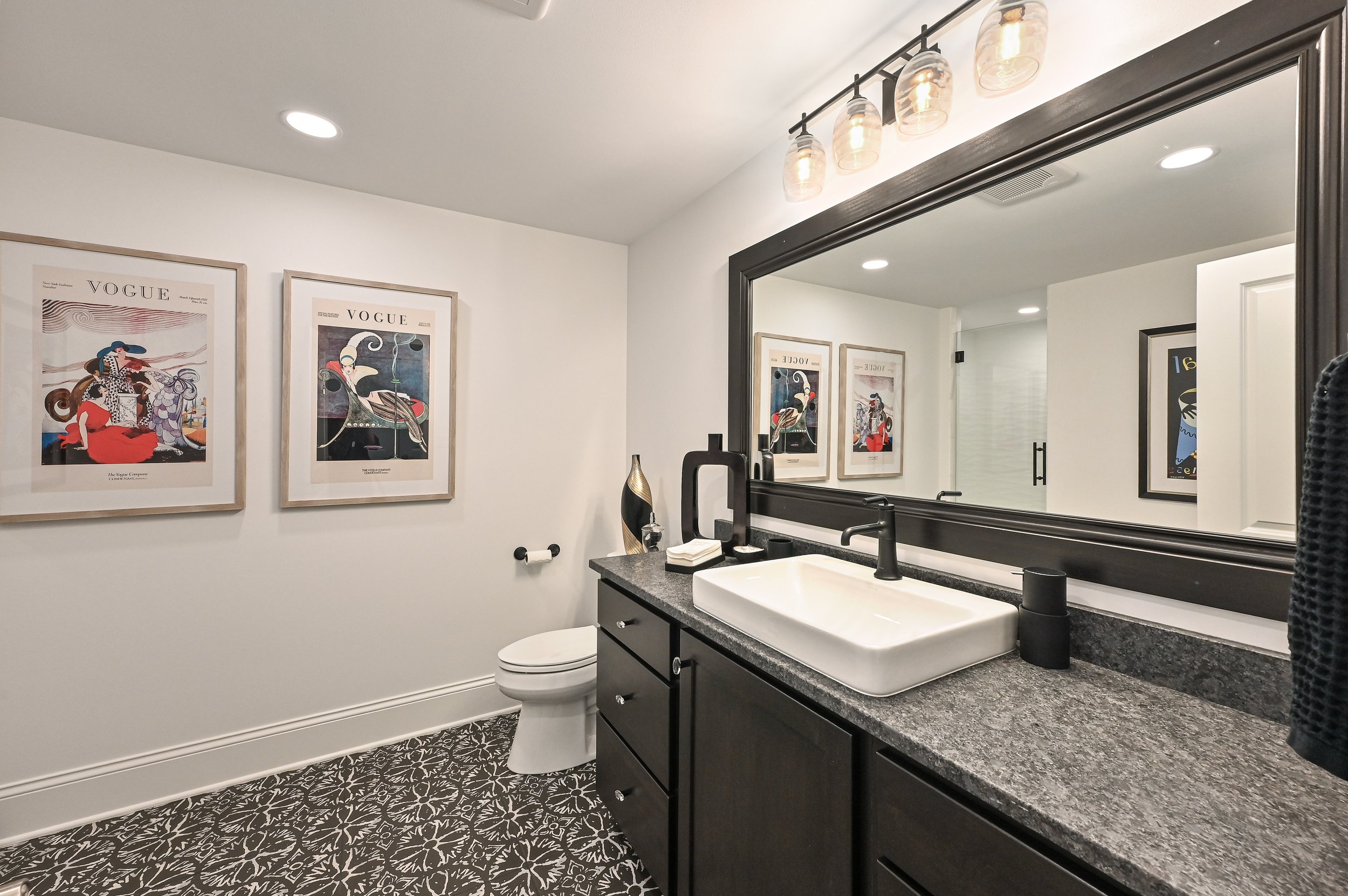 Modern bathroom with black vanity, white vessel sink, large mirror, framed Vogue magazine posters, black fixtures, patterned floor tiles, and contemporary lighting.