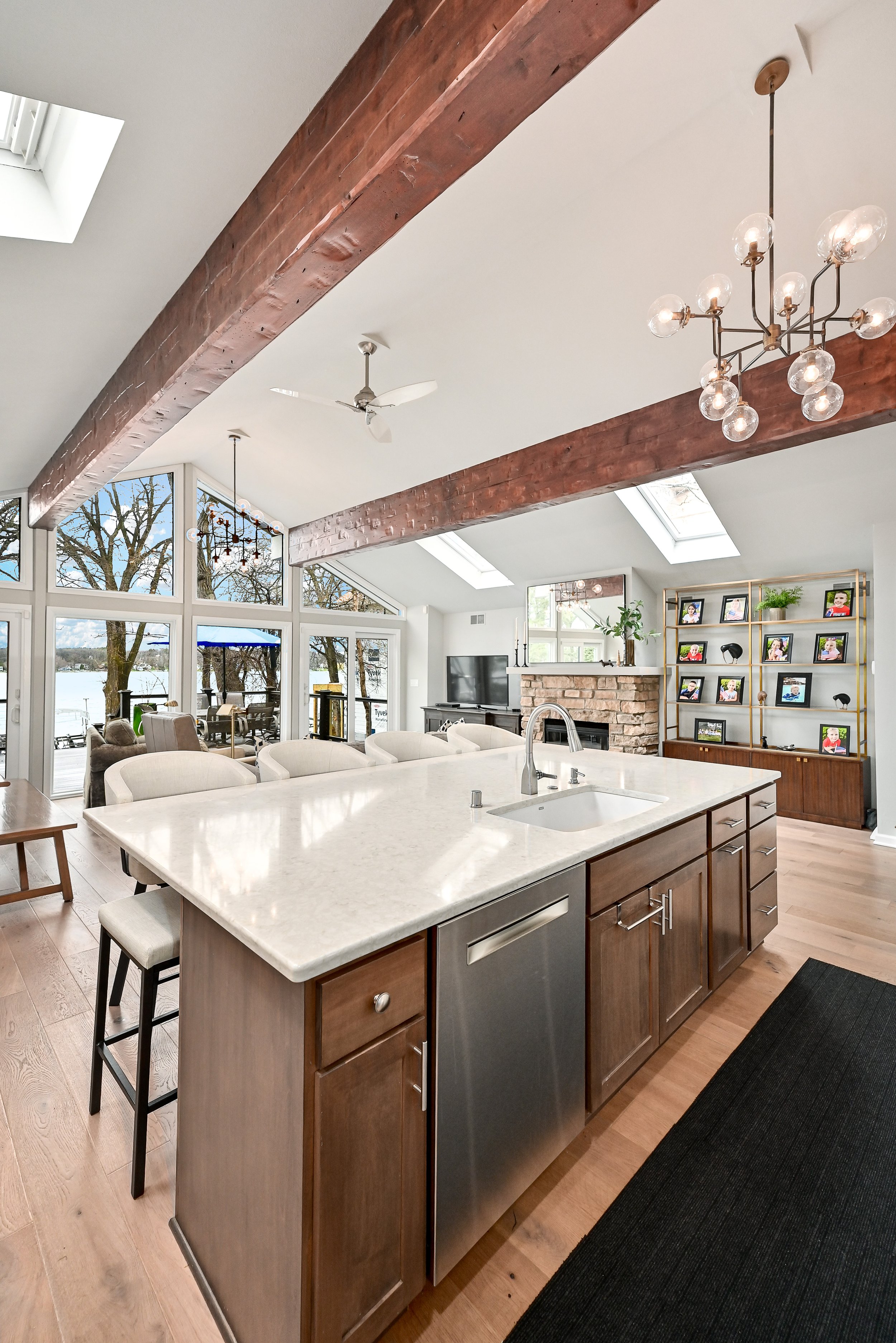 Open-concept living room and kitchen with large windows, skylights, exposed wooden beams, a stone fireplace, a TV, a shelving unit with framed photos, and modern lighting fixtures.