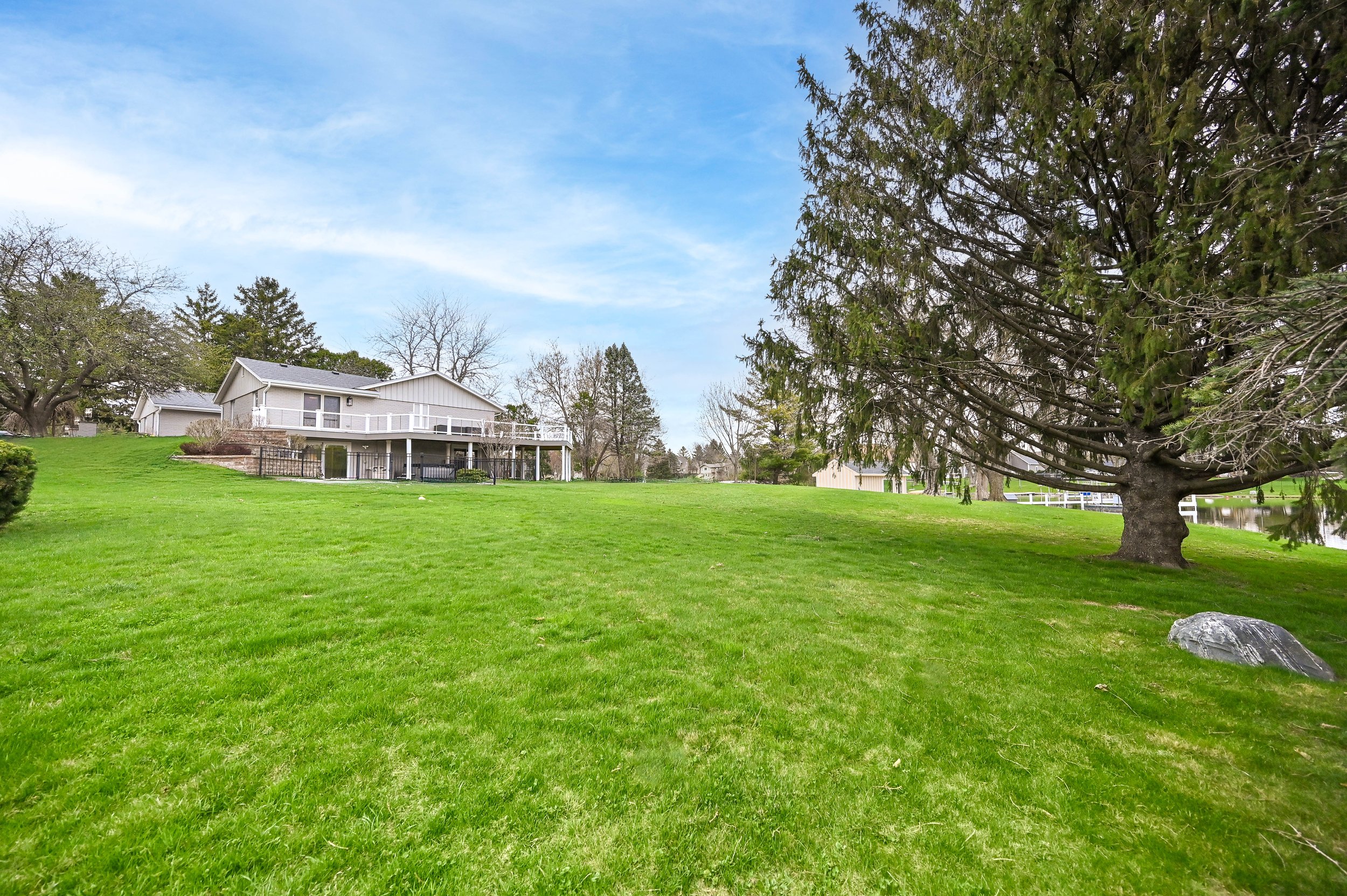 A large green grass lawn with a tree on the right and house in the background with a deck and railings, under a partly cloudy sky.