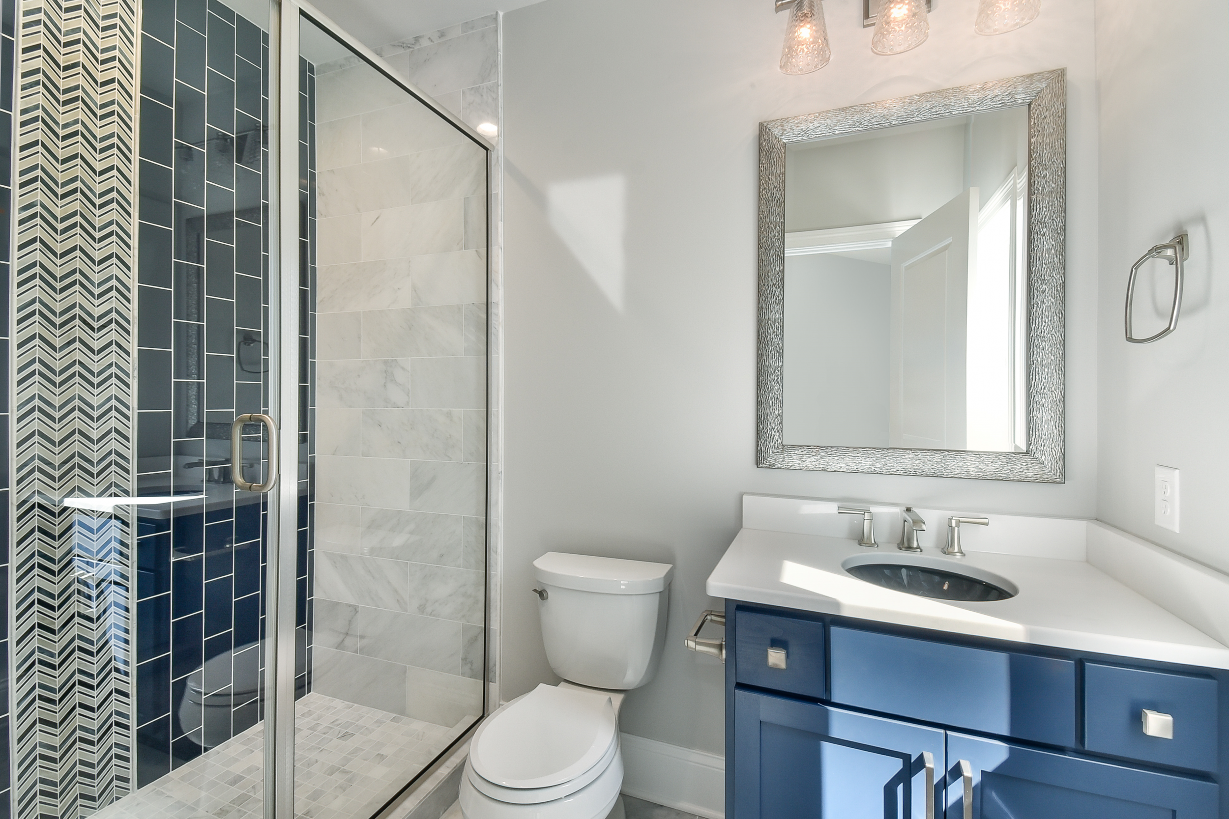 Modern bathroom with a glass-enclosed shower featuring gray and black tiles, a white toilet, a navy blue vanity with a white countertop, a black sink, a decorative mirror, and three hanging pendant lights.