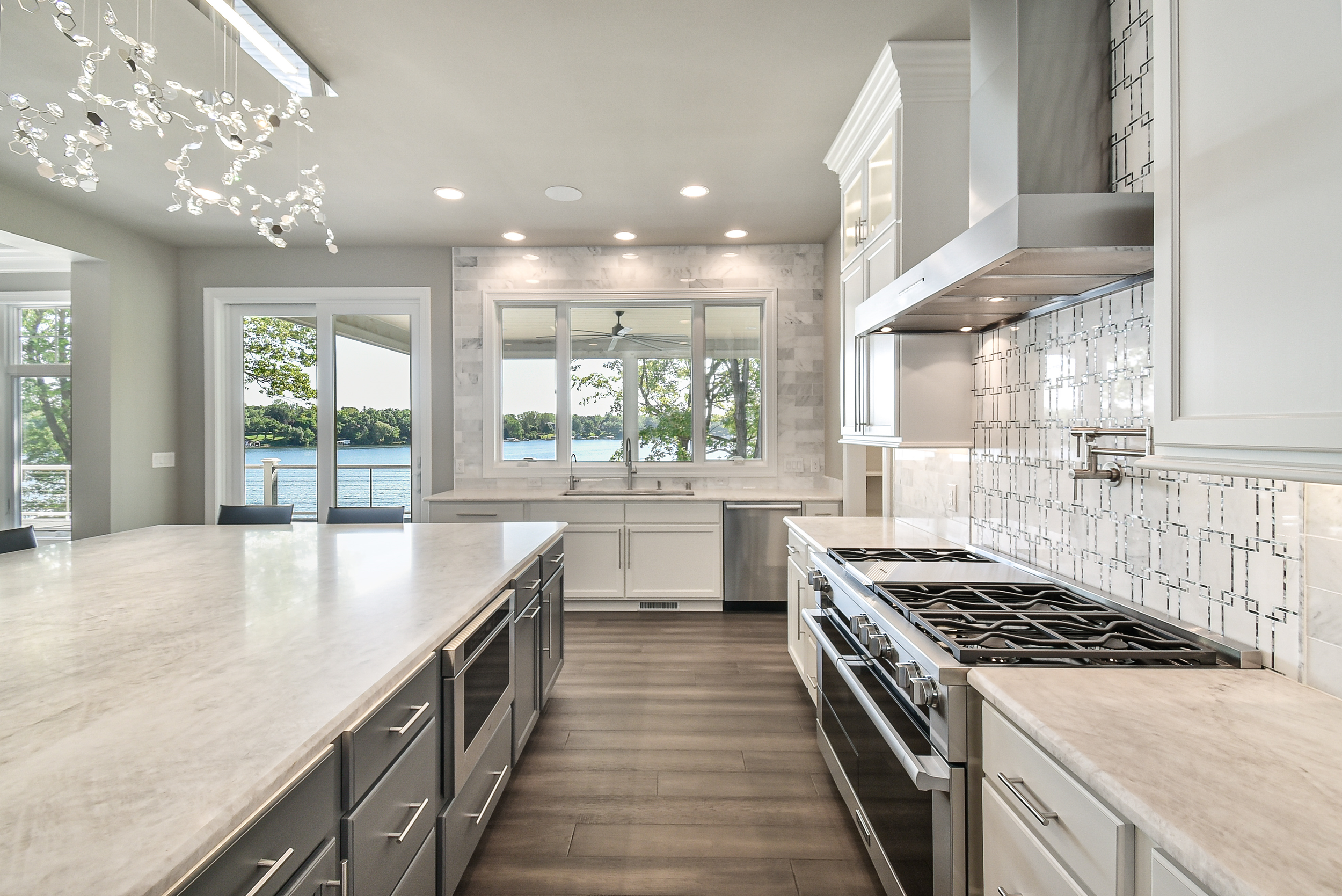 Modern kitchen with white cabinetry, marble countertops, stainless steel appliances, a large island, and a view of the lake through large windows and glass doors.