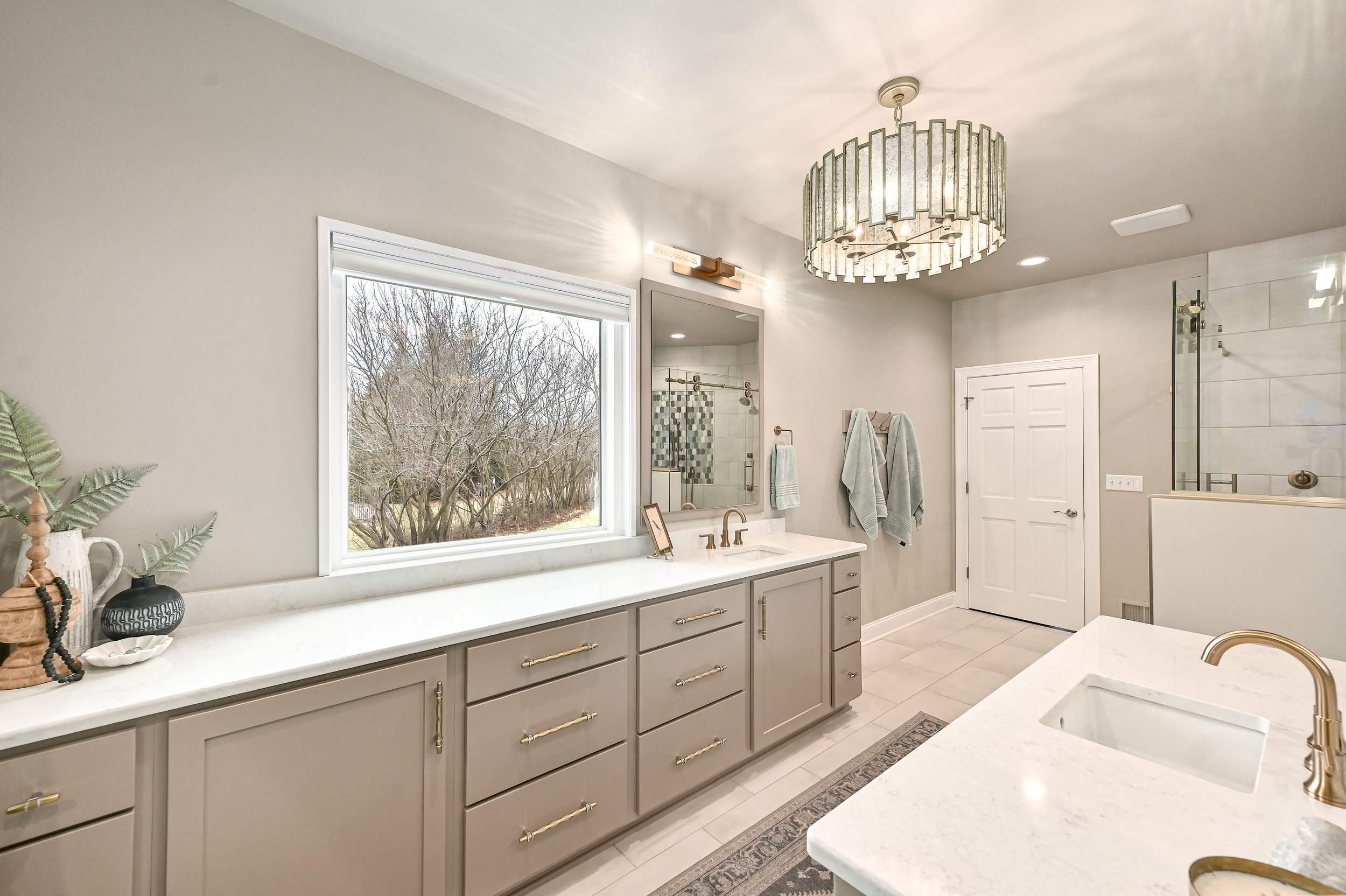 Modern bathroom with gray cabinets, white countertops, and gold fixtures. Features a window with an outdoor view, a mirror, and a hanging chandelier. Includes a shower area with glass door and a towel rack with towels.