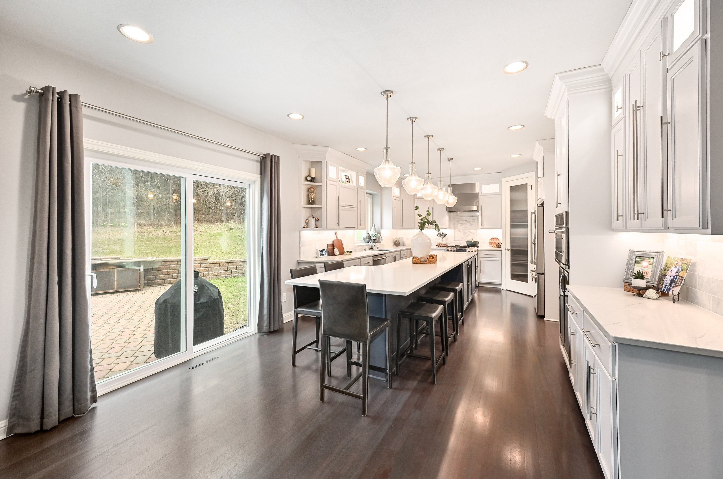Modern kitchen with white cabinets, dark wood flooring, and a large island with barstools. Sliding glass door with gray curtains leading to a backyard patio.