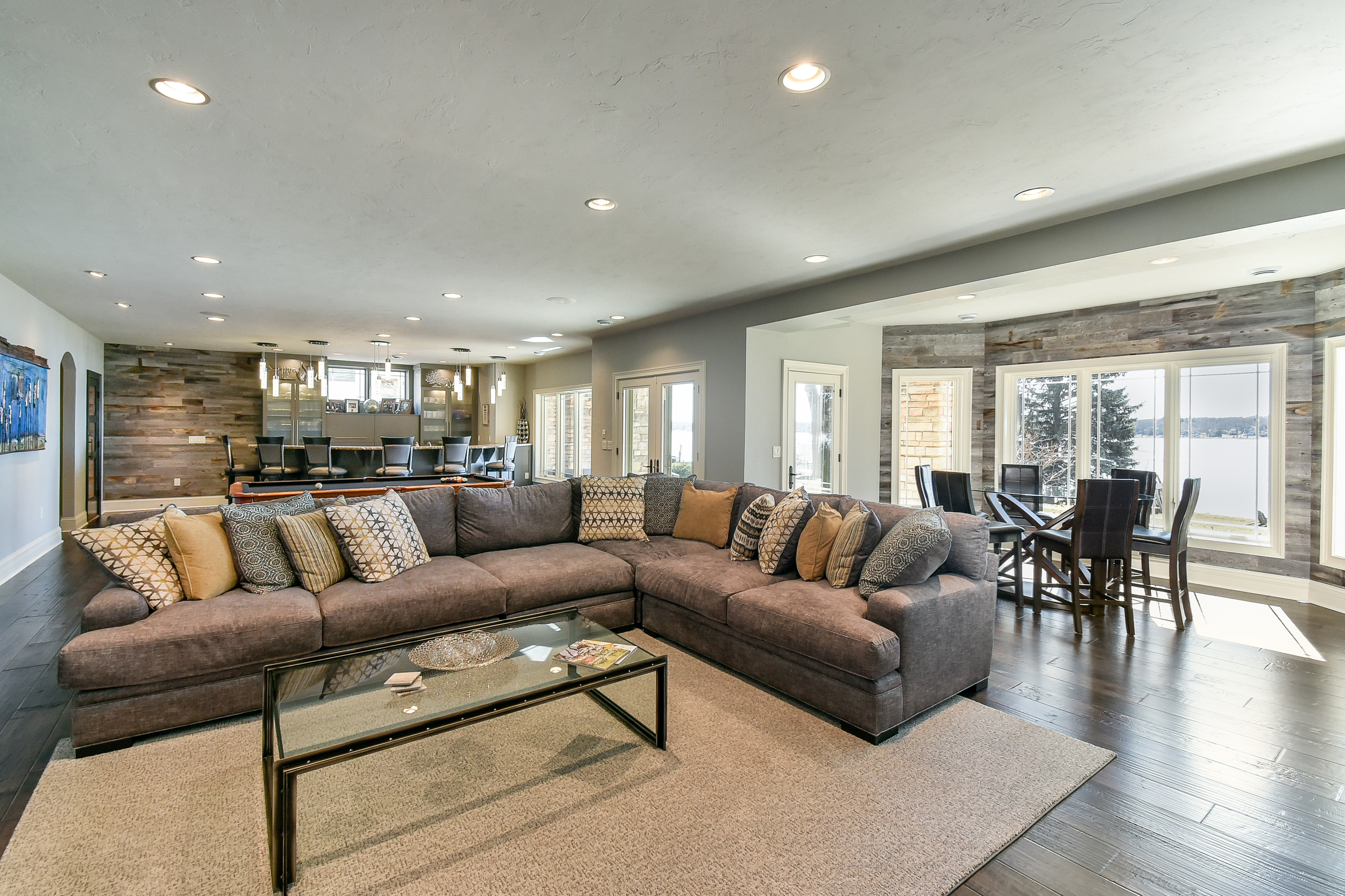 Living room with sectional sofa, decorative pillows, coffee table, wooden flooring, and large windows overlooking water and trees.