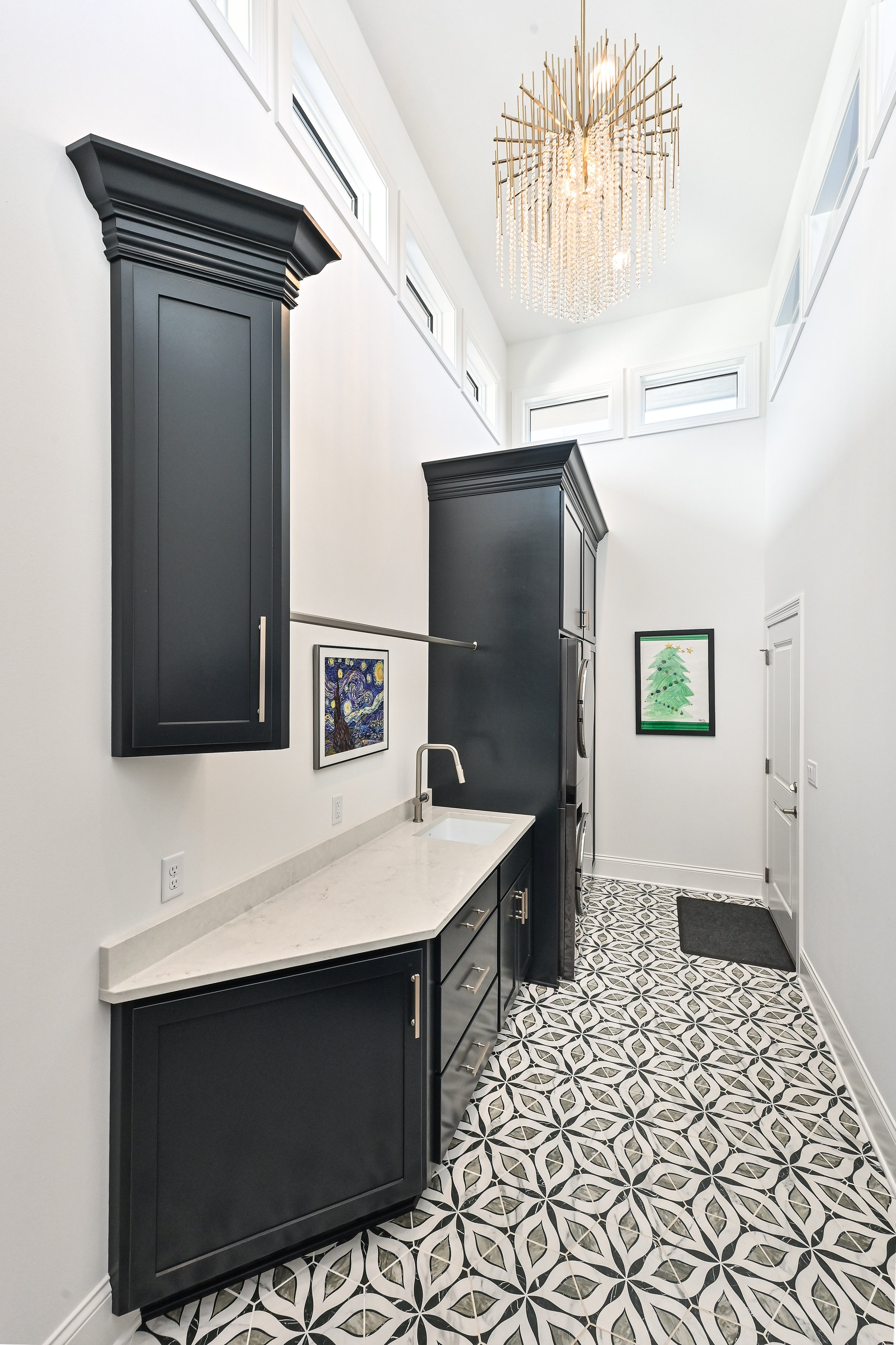 Modern laundry room with black cabinetry, white countertops, a small sink, patterned black and white floor tiles, and artwork on the walls, with high windows and a chandelier hanging from the ceiling.