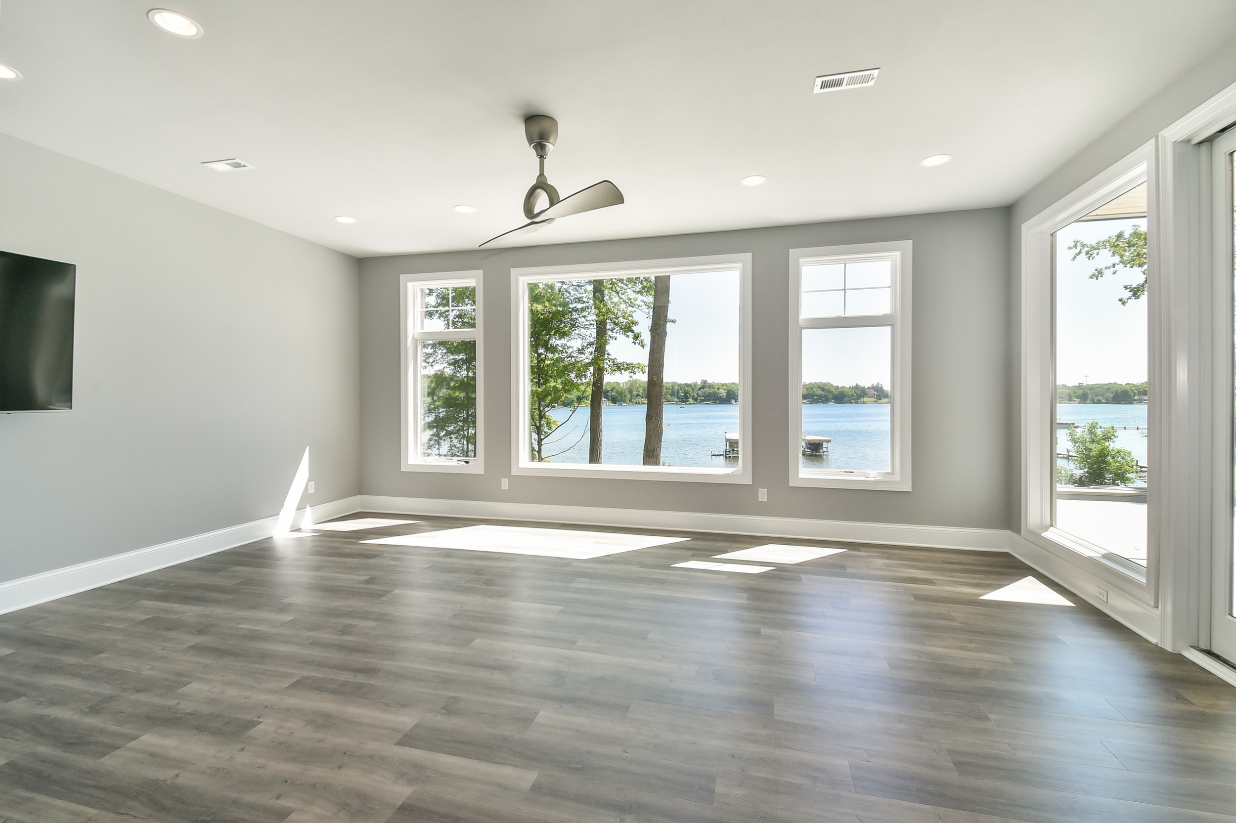 Empty living room with large windows overlooking a lake, gray walls, wood flooring, and a ceiling fan.
