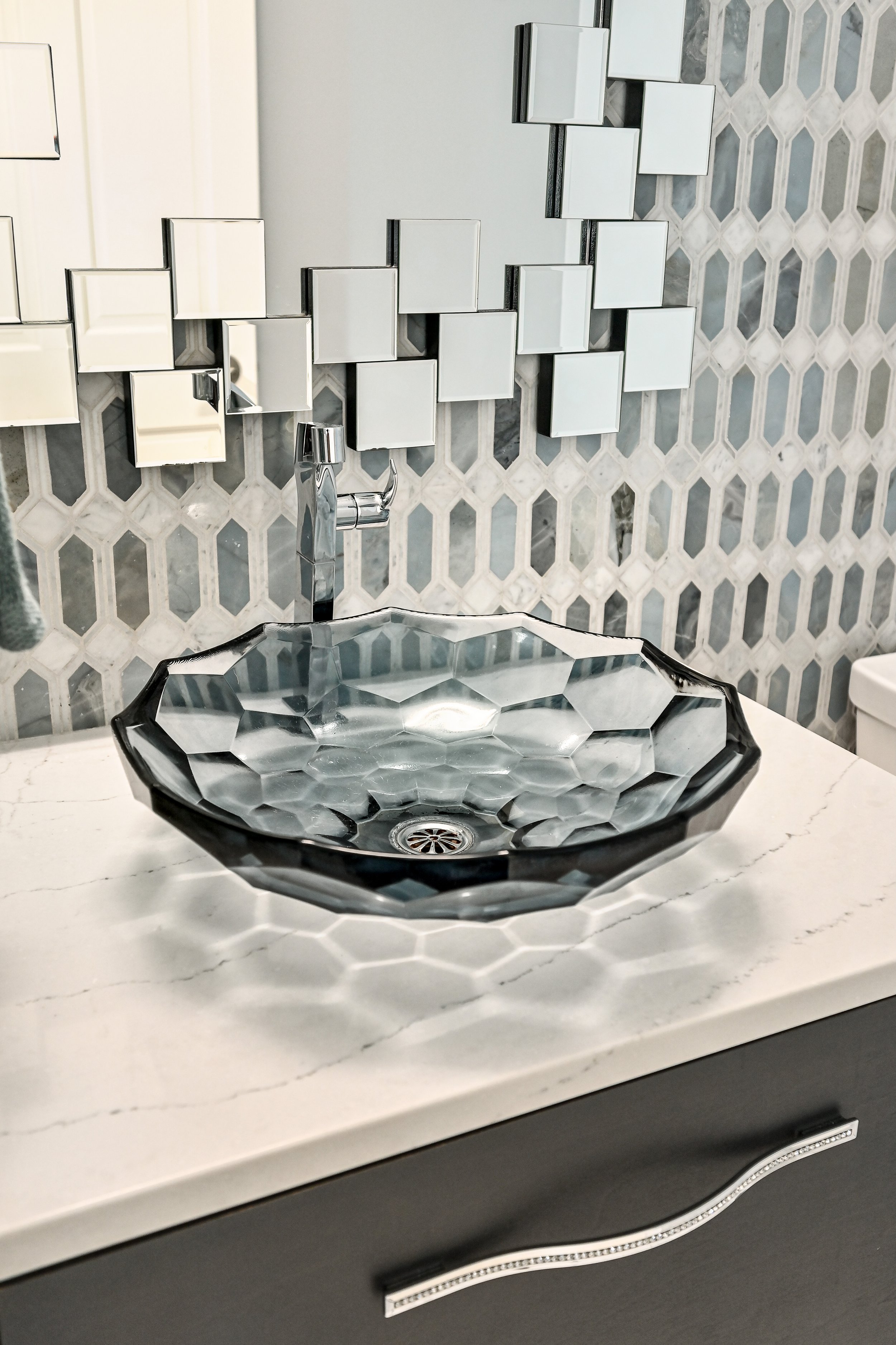 Modern bathroom sink with a geometric glass basin, decorative tile backsplash, and a mirror with assembled square tiles.