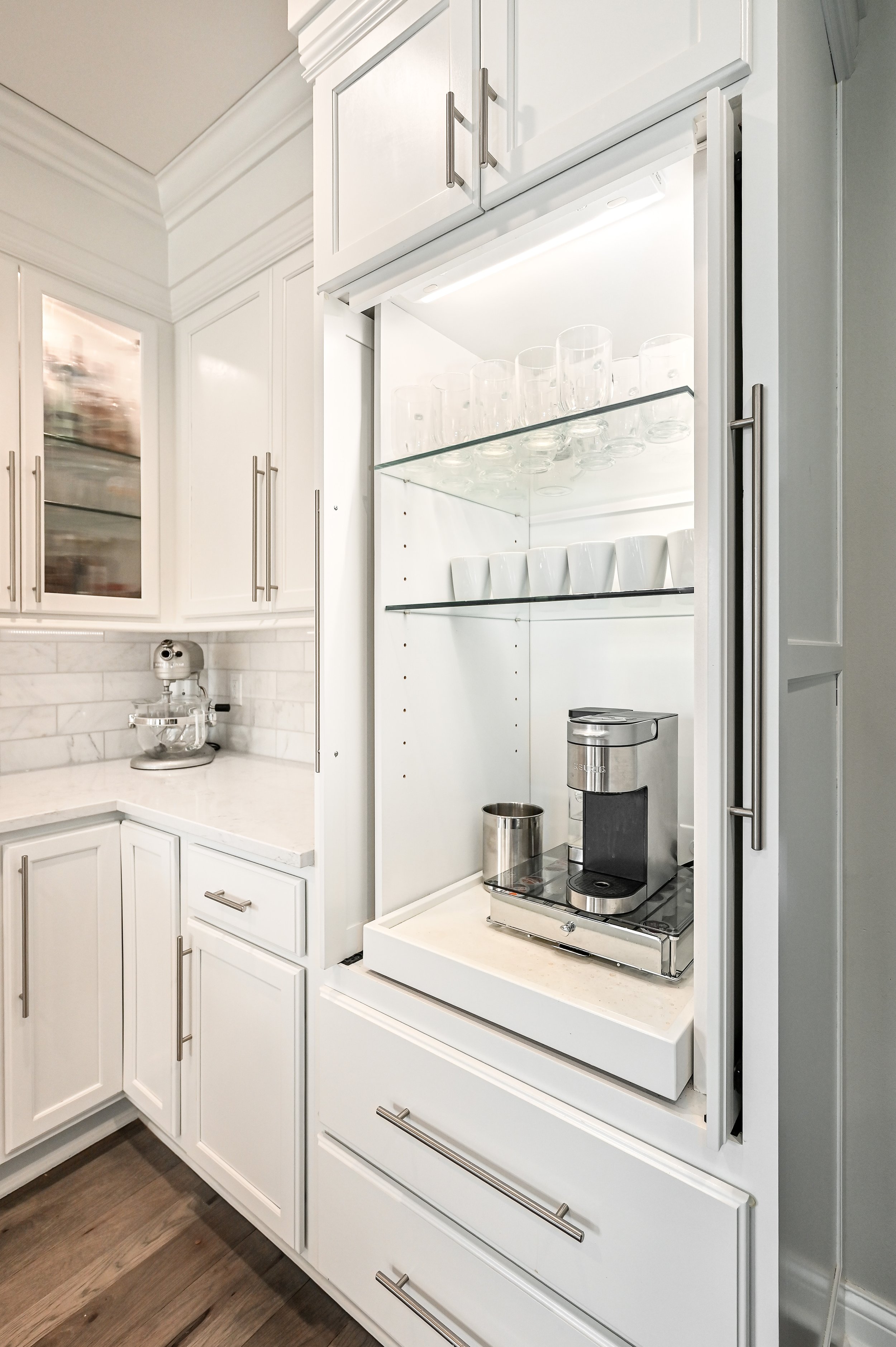 Built-in coffee station inside a white kitchen cabinet with glass shelves, a coffee machine, and stacked glasses and cups.