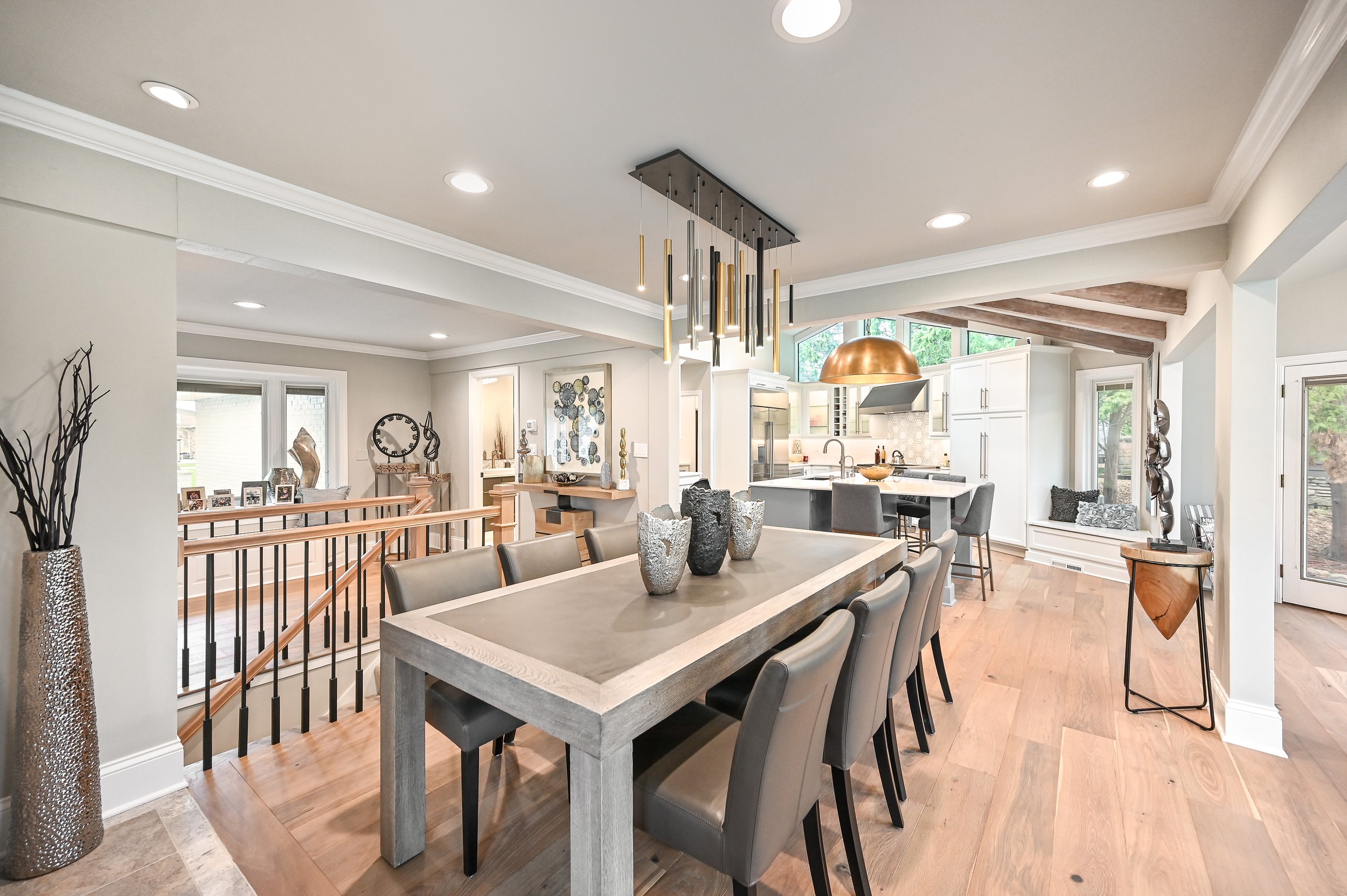 Open-concept kitchen and dining area with a large wooden dining table, gray chairs, and modern decor, illuminated by recessed lighting and a hanging chandelier.