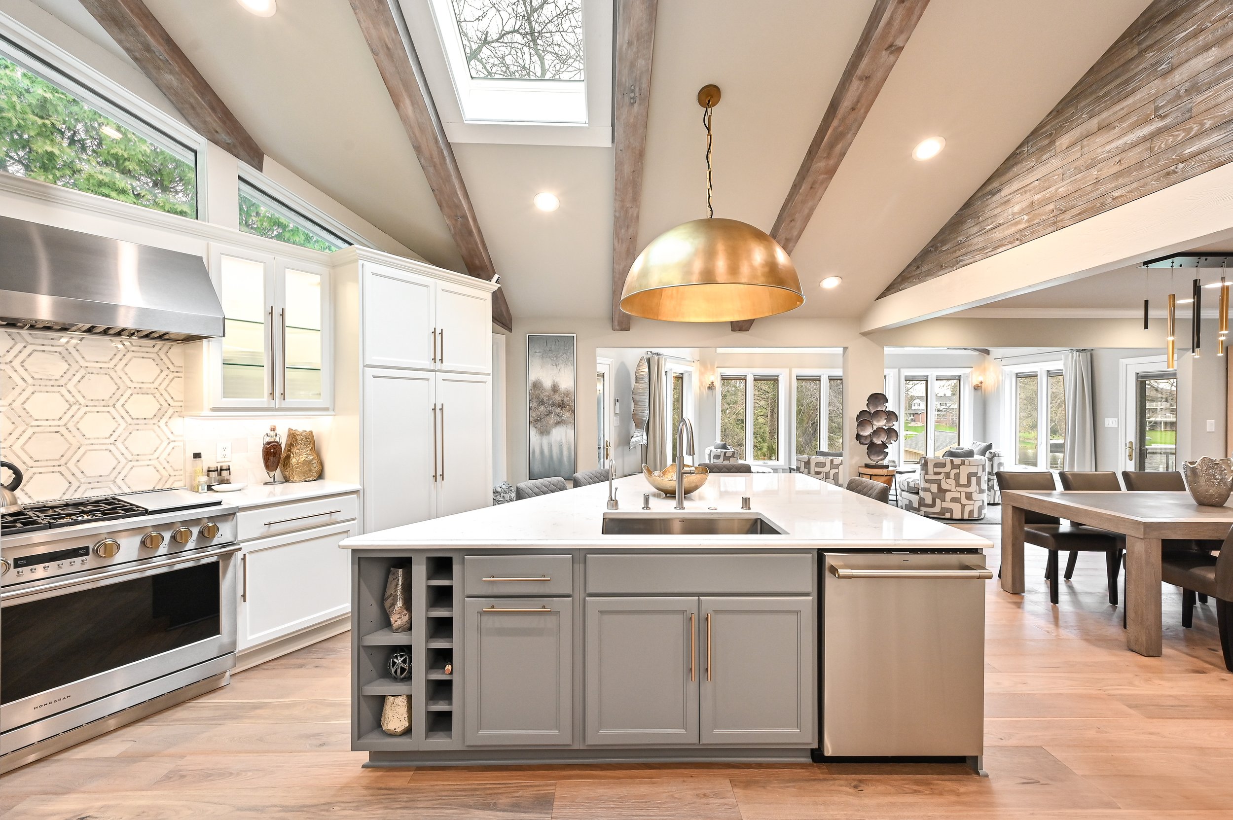 Modern open-concept kitchen with white cabinetry, stainless steel appliances, light wood flooring, a white island with storage and a sink, and a gold pendant light hanging above. In the background, there is a dining area and a living room with large 