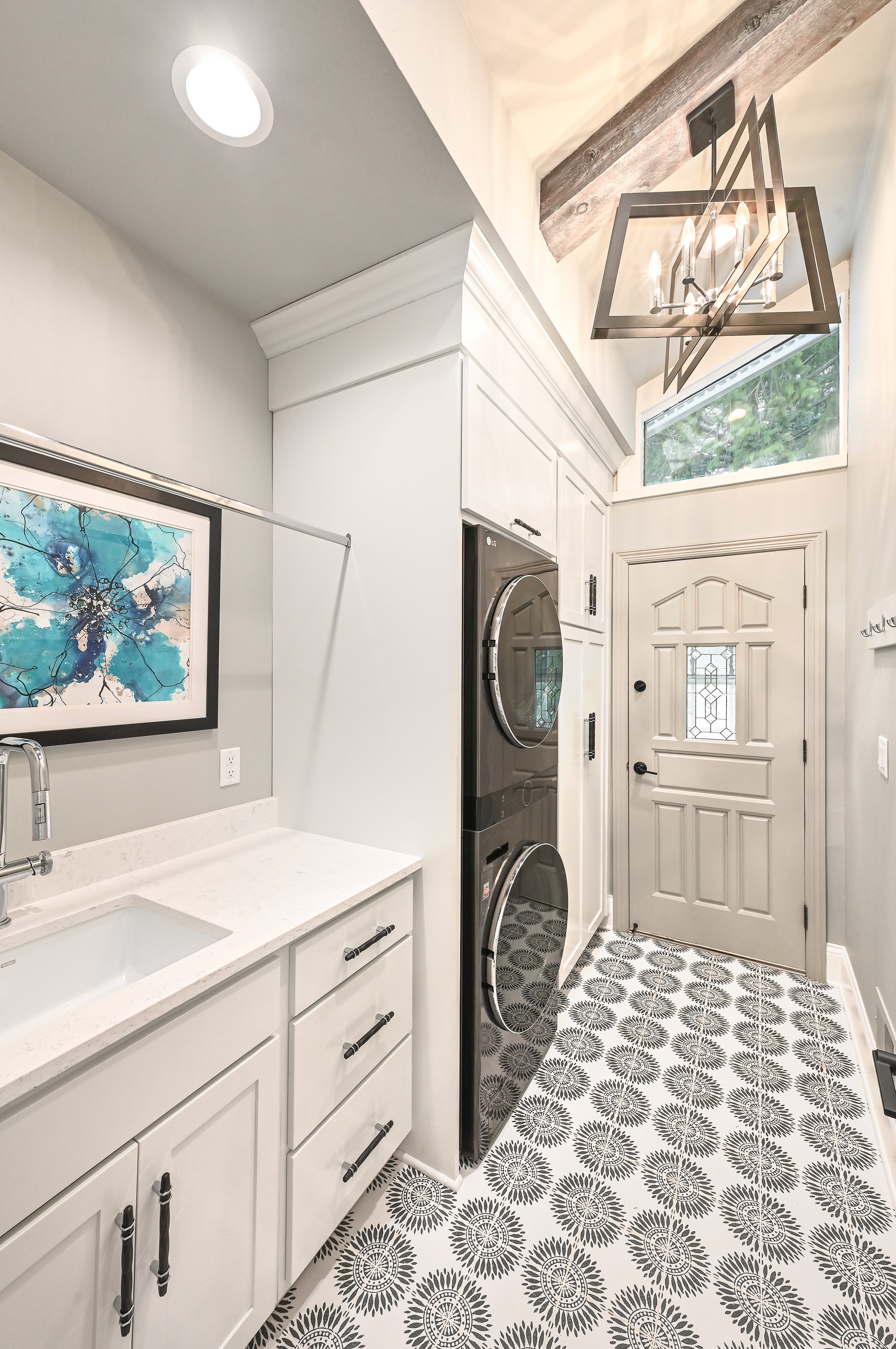 Laundry room with white cabinets, black washer and dryer stacked, patterned black and white tile floor, window, pendant light, and decorative artwork.
