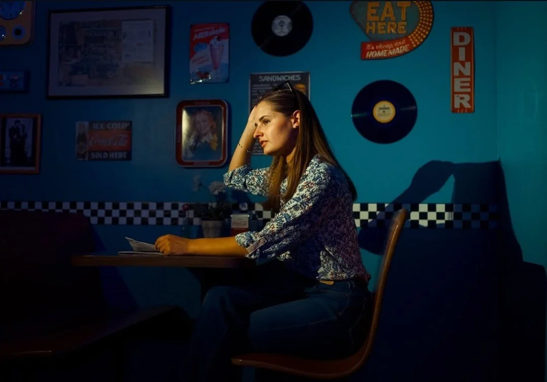 A woman sitting alone at a table in a dimly lit restaurant with retro decor, including vinyl records on the wall and vintage signs. Carolyn Kashmanian, Jazz, Live music, Charlotte, North Carolina, bebop, singers, vocalists, eighty eights, Petra's