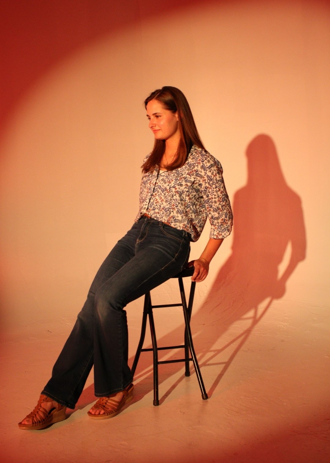 A woman with brown hair sitting on a black stool, wearing a floral blouse, dark jeans, and brown sandals, with a shadow cast on a plain wall behind her. Carolyn Kashmanian, Jazz, Live music, Charlotte, North Carolina, bebop, singers, vocalists, eight