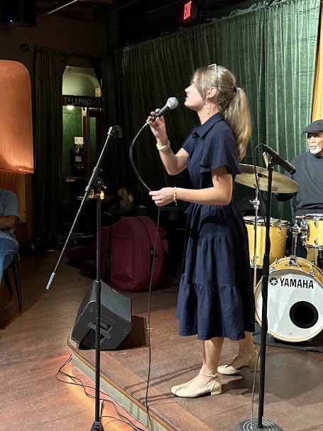 A woman with a ponytail in a dark dress singing into a microphone on small stage with a drummer playing in the background, green curtain behind, and audience members nearby. Carolyn Kashmanian, Jazz, Live music, Charlotte, North Carolina, bebop, sing